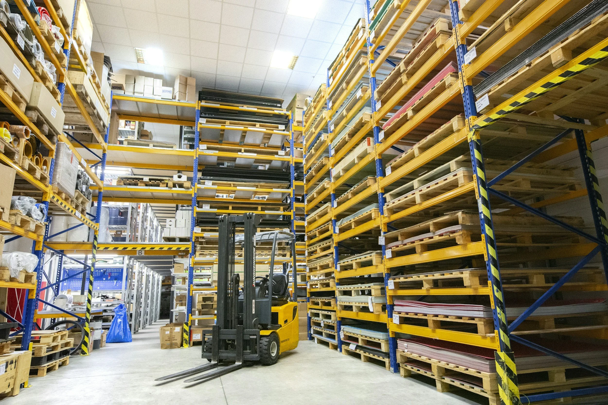 Inside a warehouse with tall yellow and blue shelving units filled with boxes, pallets, and supplies. A forklift is in the middle of the aisle, and there are additional storage racks and boxes visible in the background.