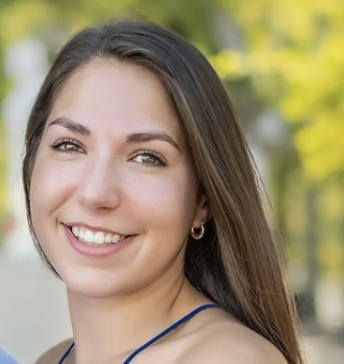 Close-up of a young woman with long brown hair smiling outdoors, with a blurred green background.