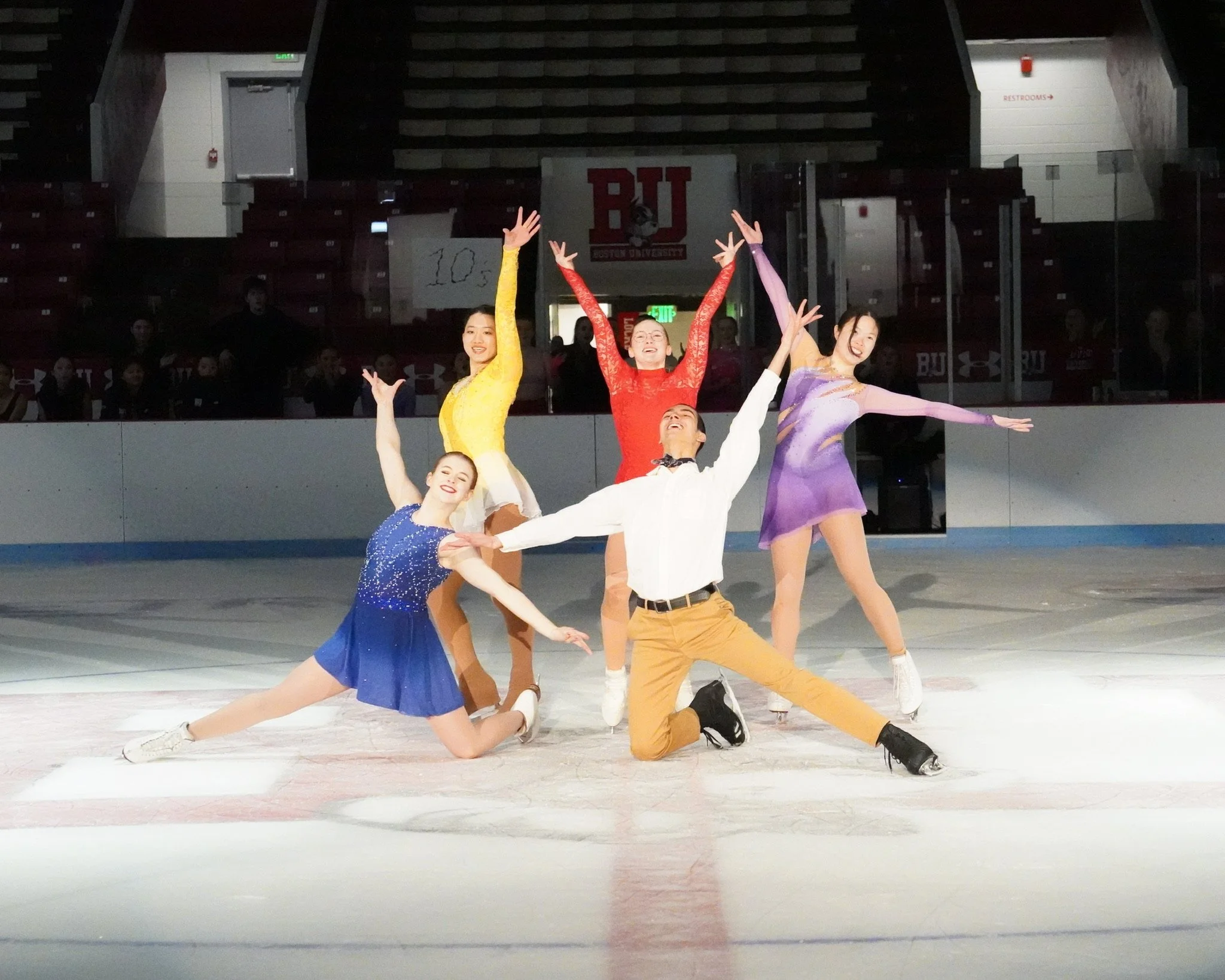 Five figure skaters in colorful costumes performing a synchronized routine on an ice rink.