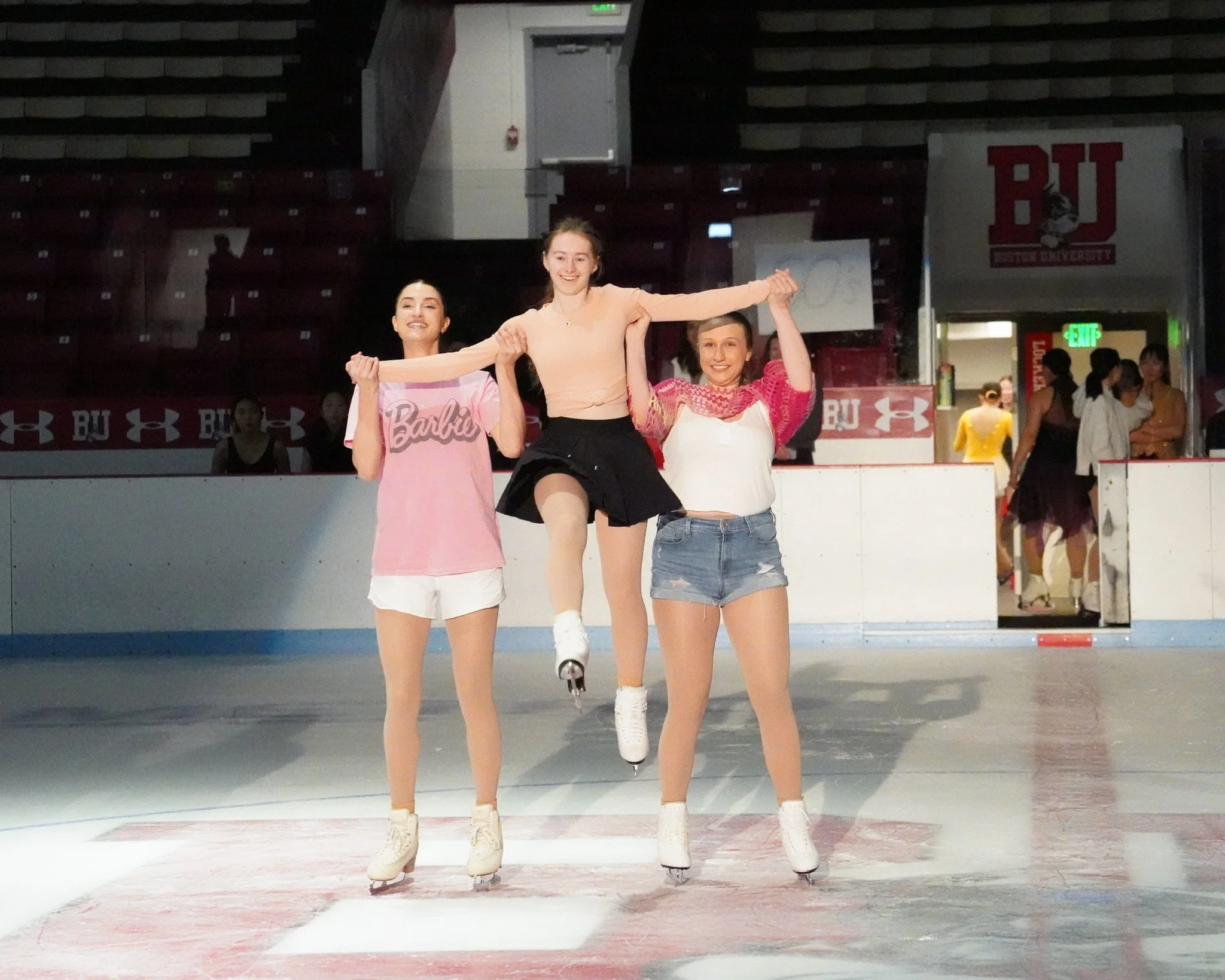 Three young women ice skating on an ice rink, with two supporting a third who is skating in the middle, holding hands in a celebratory pose, with a BU (Boston University) banner in the background.