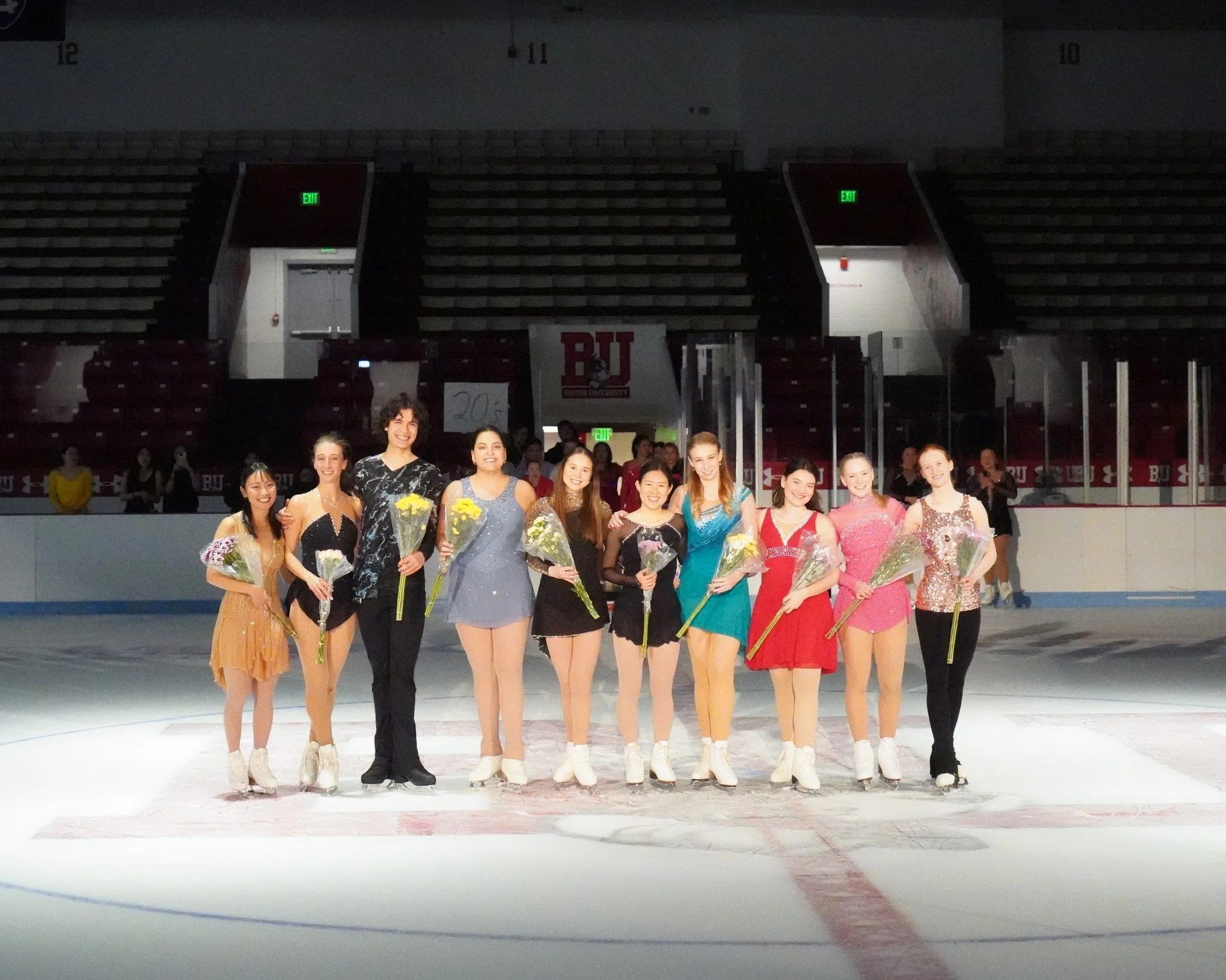 A group of ten female figure skaters in colorful costumes holding flowers, posing on an ice rink after a performance.