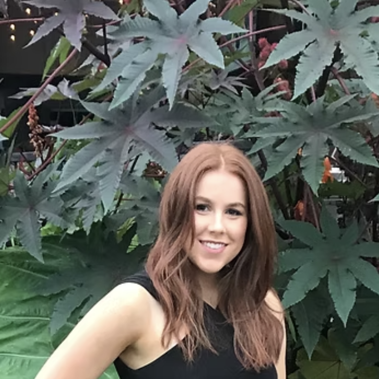 A young woman with long, wavy, auburn hair smiles at the camera in front of large green and dark red tropical leaves.