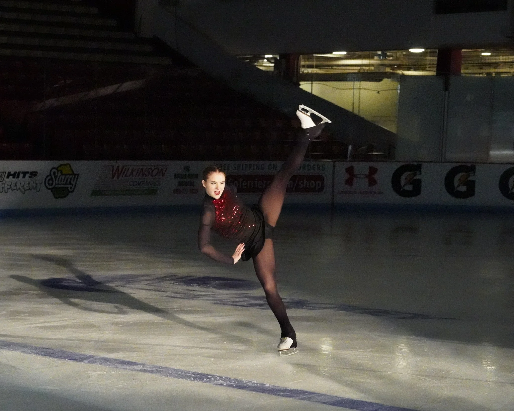 A female figure skater performing a move on the ice rink, wearing a black and red costume, with one leg raised high and one arm extended, illuminated by bright lights, with advertisements and empty stands in the background.