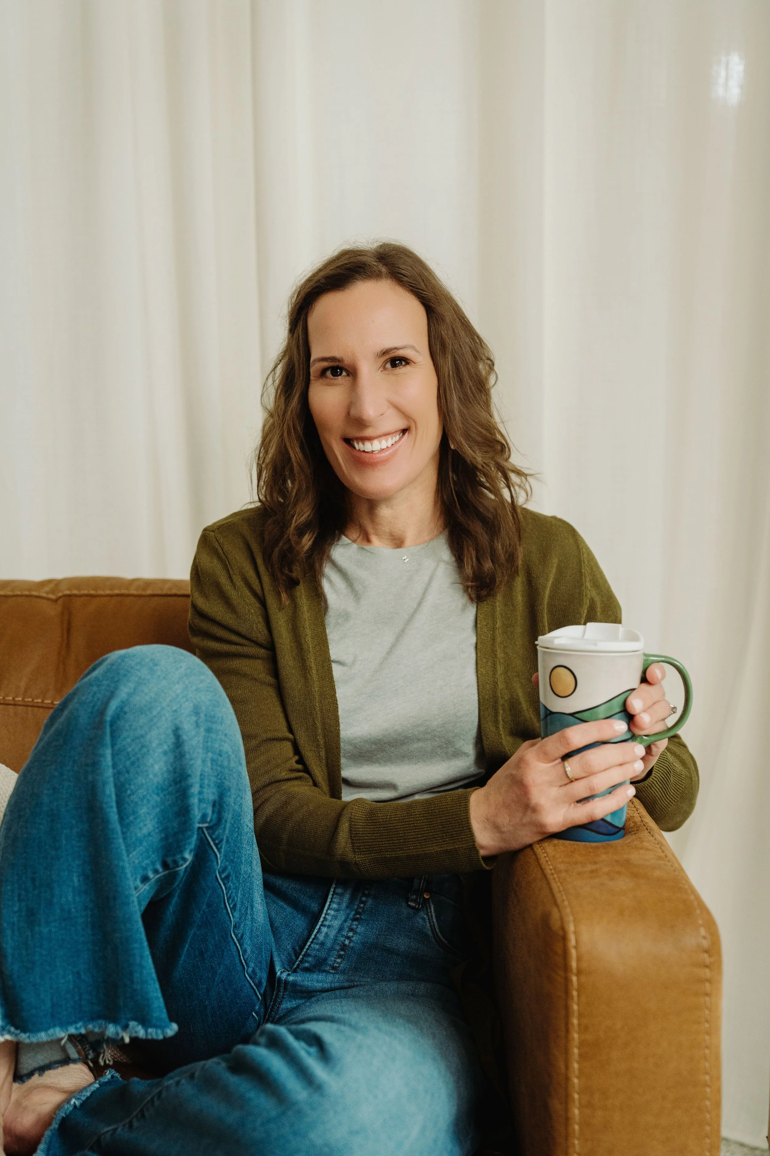 Smiling woman sitting on a couch holding a colorful mug, wearing a light gray shirt and an olive green cardigan, with a white curtain background.
