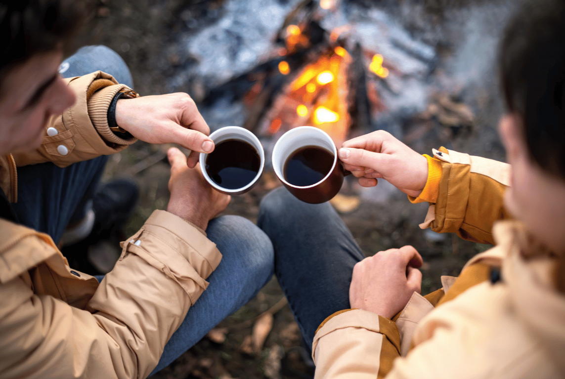 Two people sitting outdoors in front of a campfire, holding cups of coffee or tea.