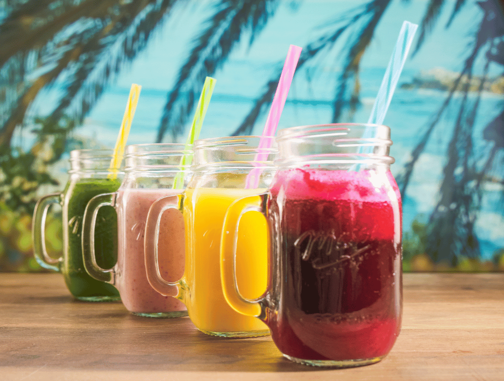 Four colorful smoothies in mason jars with straws on a wooden surface against a tropical beach background.