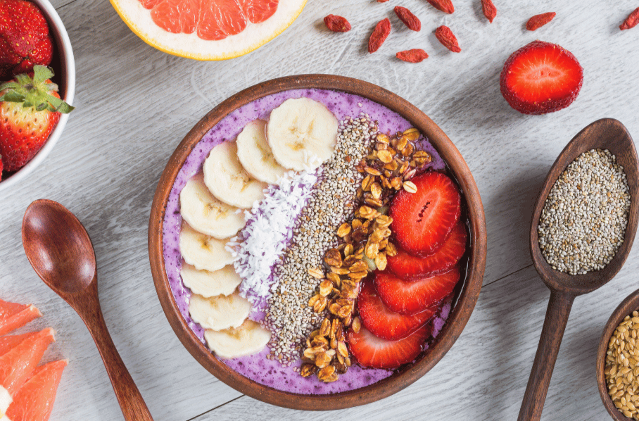 A bowl of purple smoothie topped with sliced bananas, strawberries, shredded coconut, chia seeds, and granola, surrounded by fresh fruit and scattered goji berries.