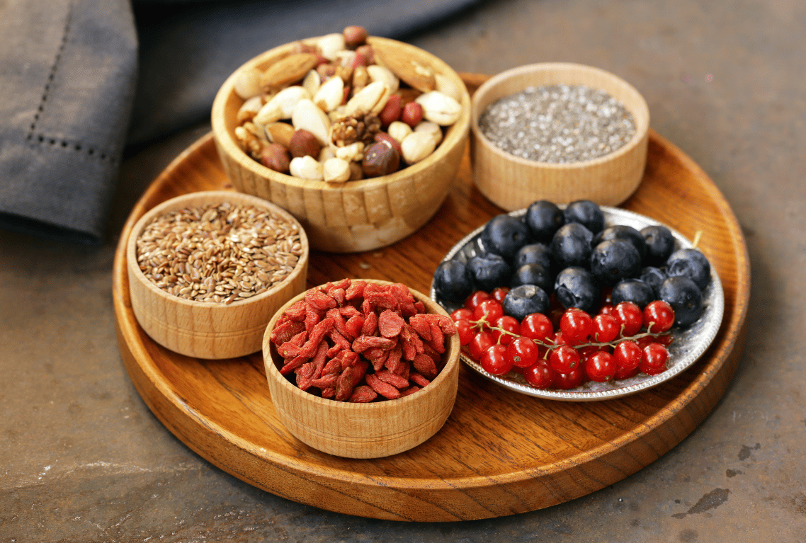 Assorted seeds, berries, and nuts on a wooden tray, including blueberries, red currants, goji berries, chia seeds, flax seeds, and mixed nuts.
