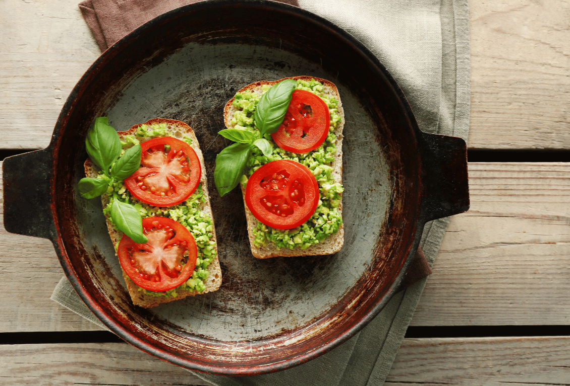 Two slices of bread topped with mashed avocado, tomato slices, and fresh basil leaves on a black skillet.