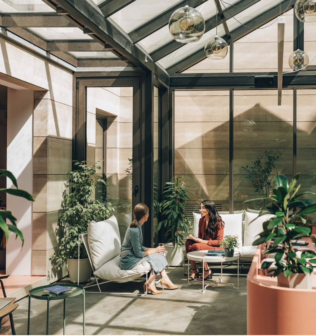 Deux personnes discutent dans un salon lumineux avec beaucoup de plantes, des fauteuils blancs et une grande fenêtre en verre avec une pleine vue vers l'extérieur.