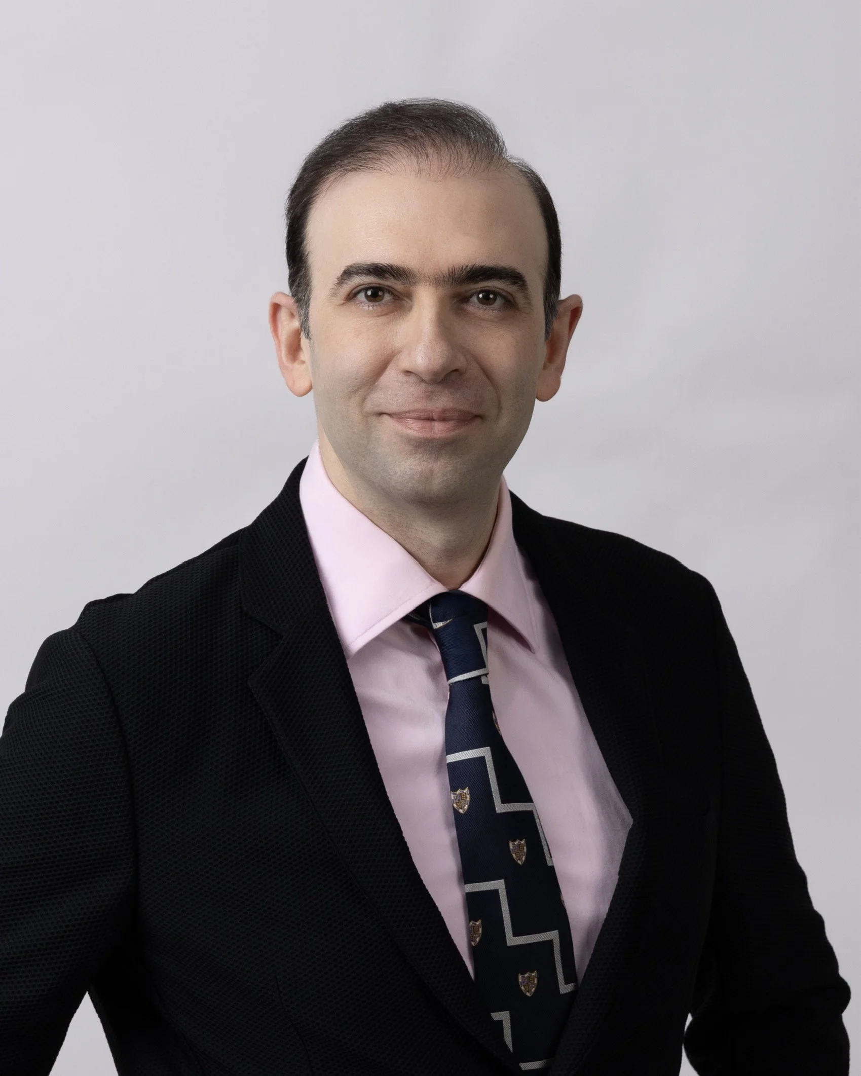 A man dressed in a black suit, pink shirt, and a patterned tie, smiling at the camera with a plain light gray background.