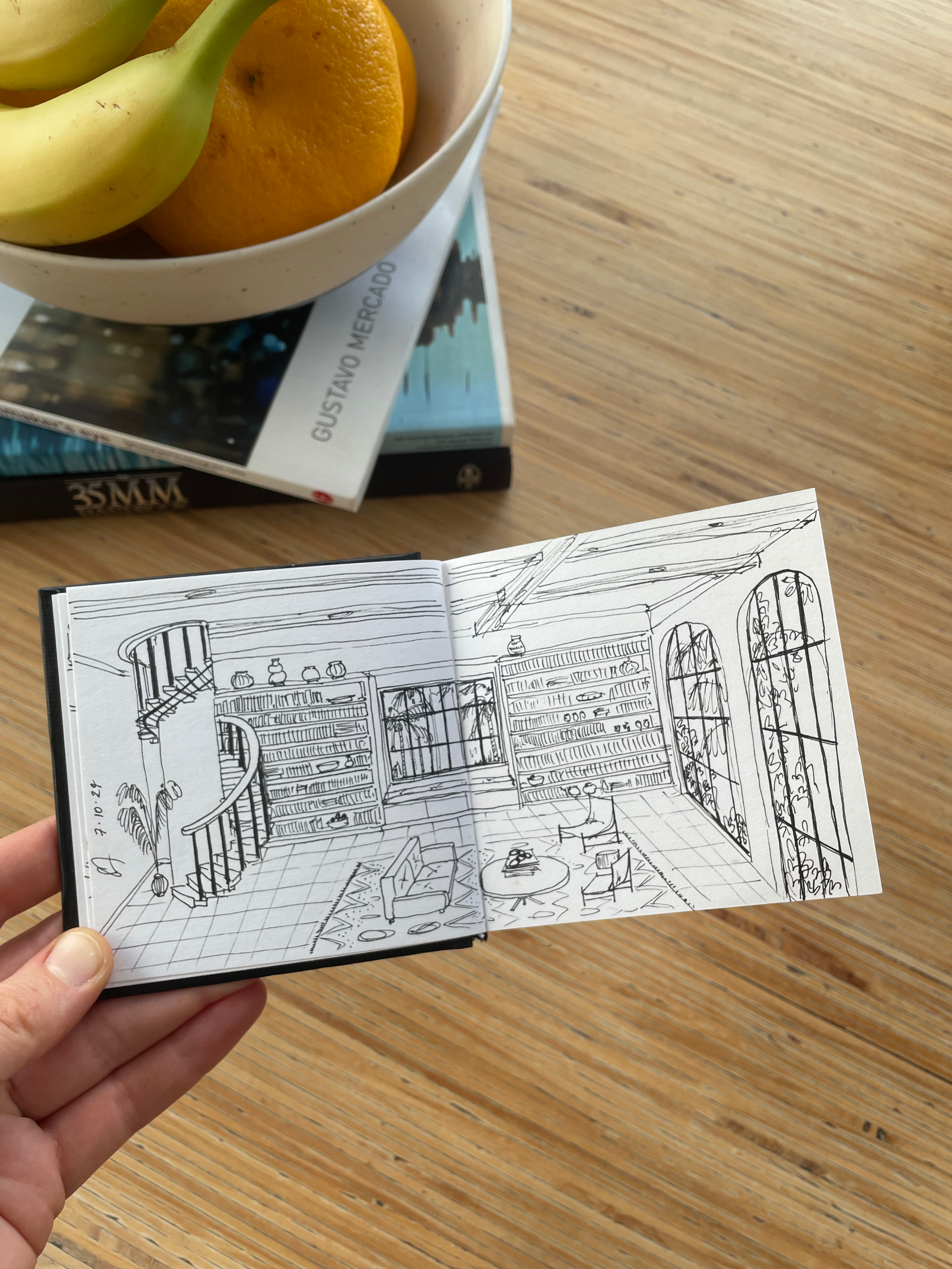Photo of a person's hand holding a sketchbook with a detailed interior room drawing, on a wooden surface with candles, books, and a bowl of fruit in the background.