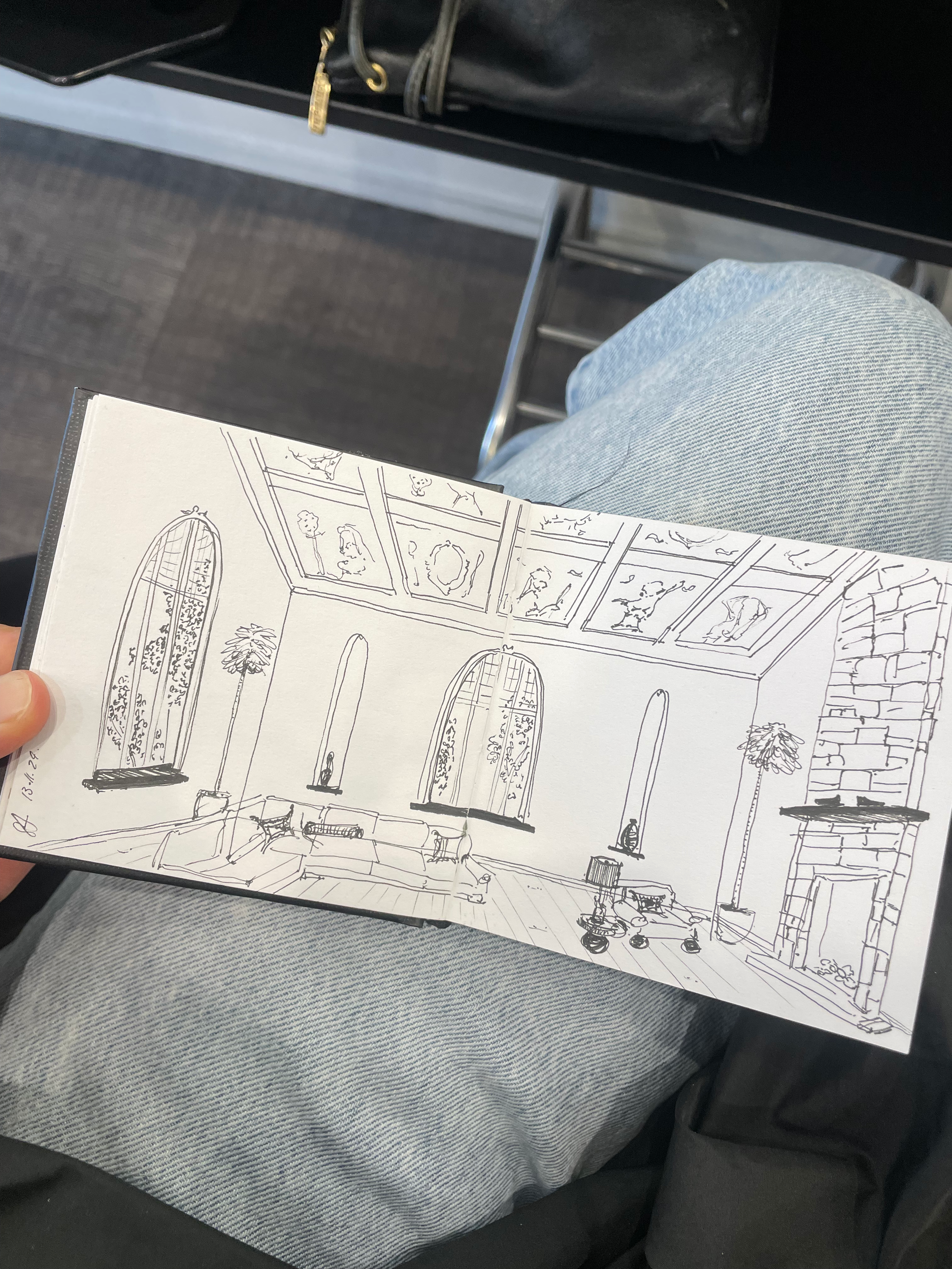 A hand holding a sketch of an interior room with tall arched windows, plants, and a ceiling with artwork, while sitting in a blue jeans. Part of a black table or shelf and black bag are visible.