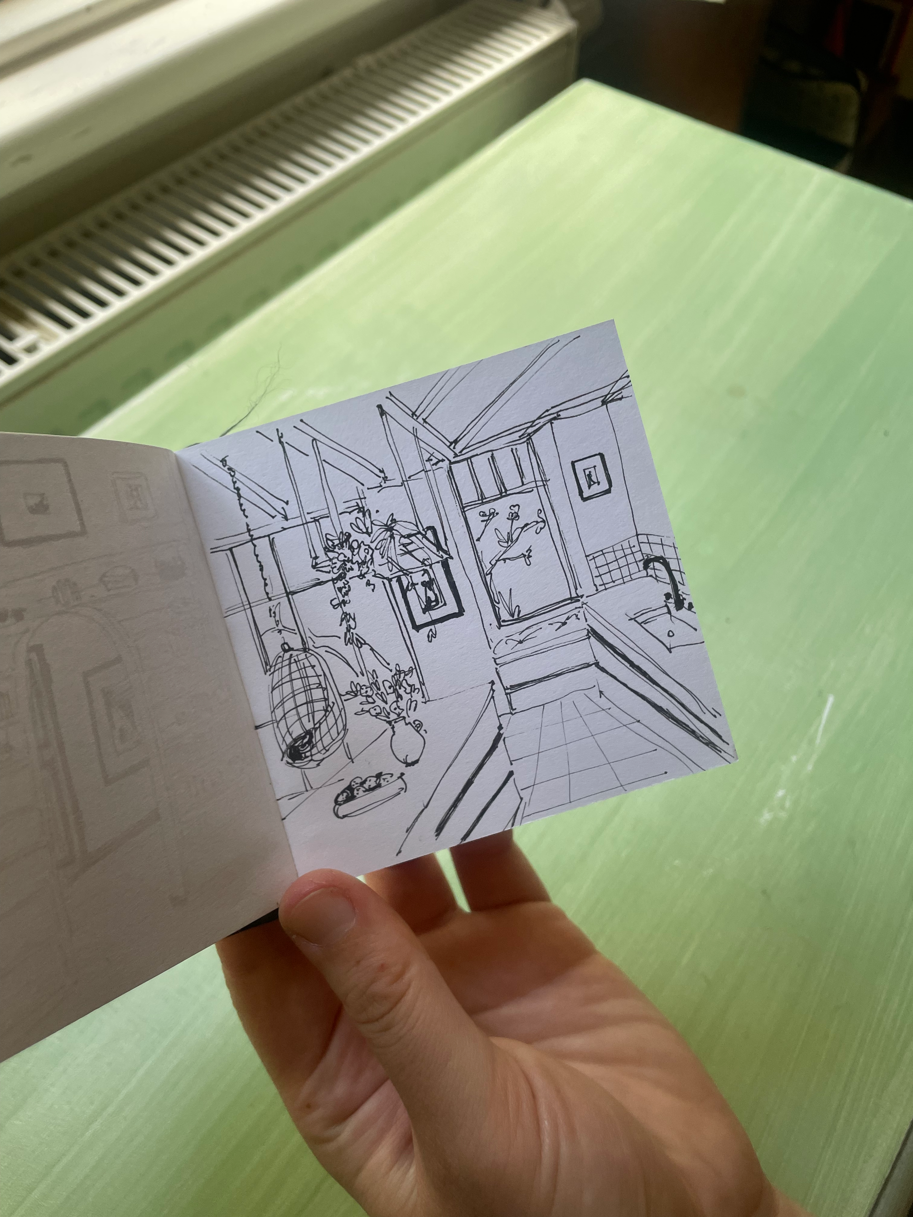 A hand holding a small sketch of a kitchen interior with plants, artwork, a window, and a tiled floor.