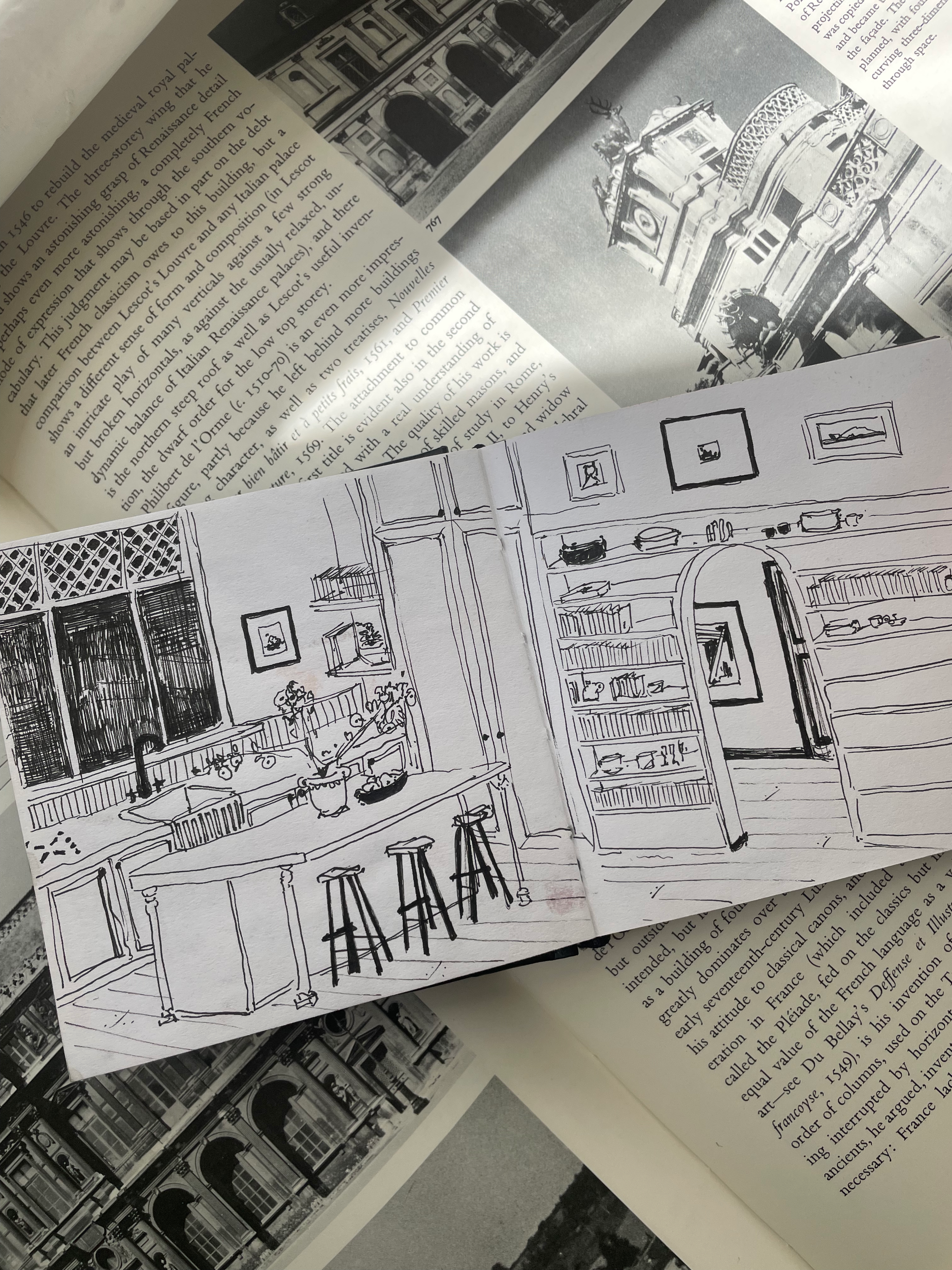 A sketch of a kitchen interior featuring an island with three stools, cabinets, shelves with decorative objects and framed pictures on the wall.