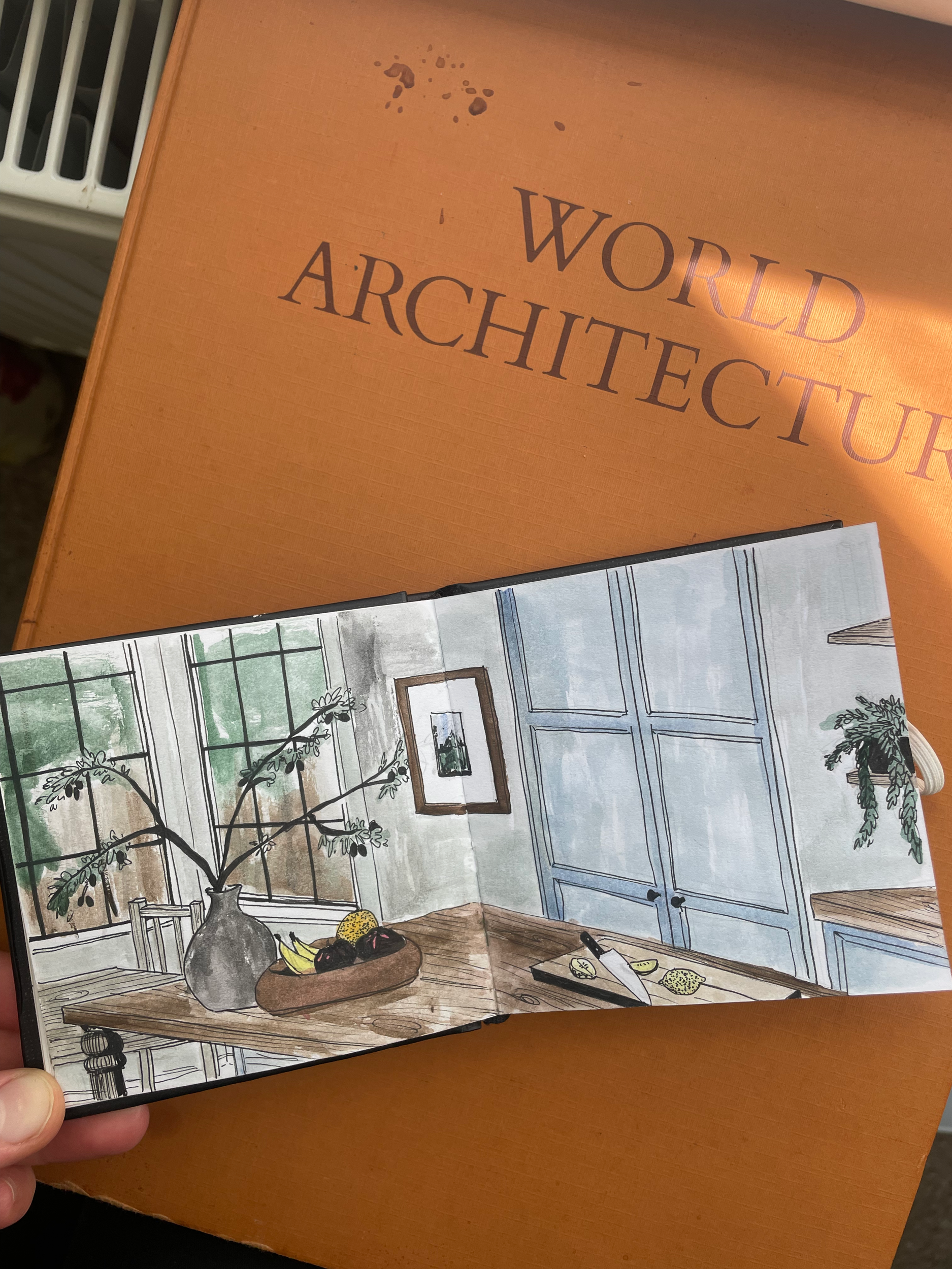 An open illustrated booklet showing a dining room with a wooden table, a gray vase with branches, a fruit bowl, and framed wall art, in front of a closed cabinet with blue doors, with a window in the background.