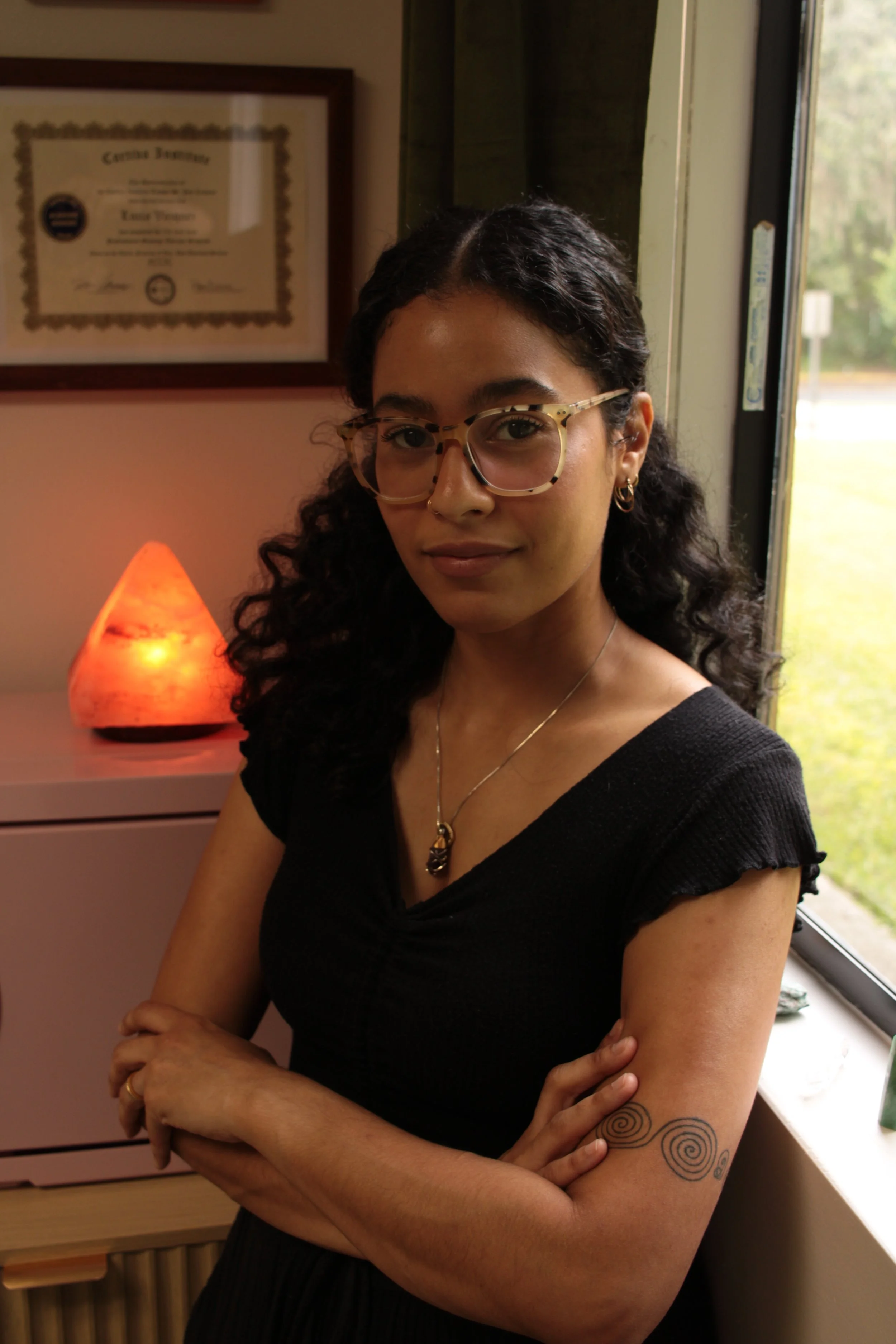 A woman with glasses and curly hair standing indoors near a window, with a salt lamp and framed certificate in the background.