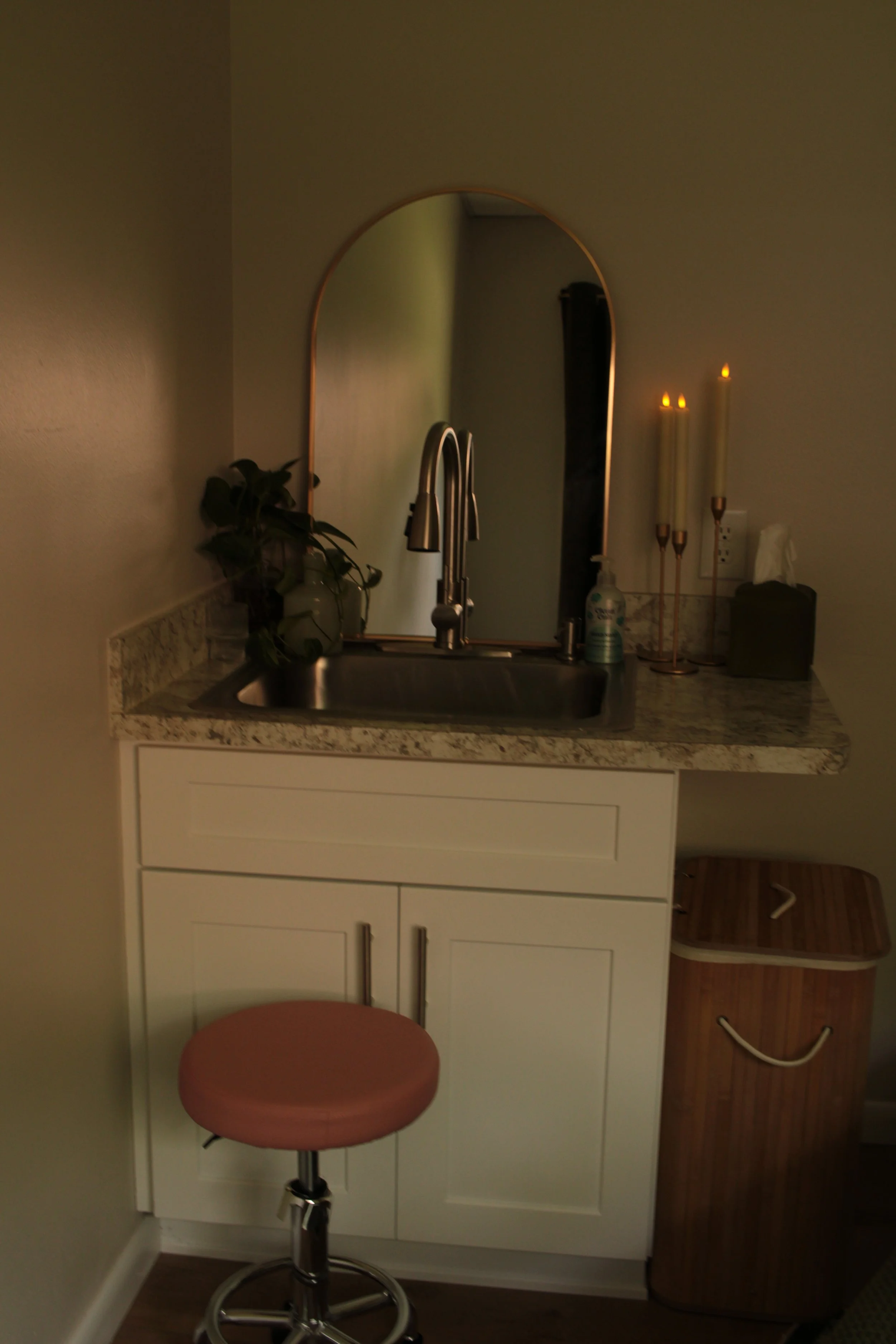 A small kitchenette with a white cabinet, a granite countertop, a stainless steel sink, a mirror, two lit candles, a plant, hand sanitizer, and a tissue box. A pink office stool is in front of the cabinet, and a brown laundry basket is to the right.