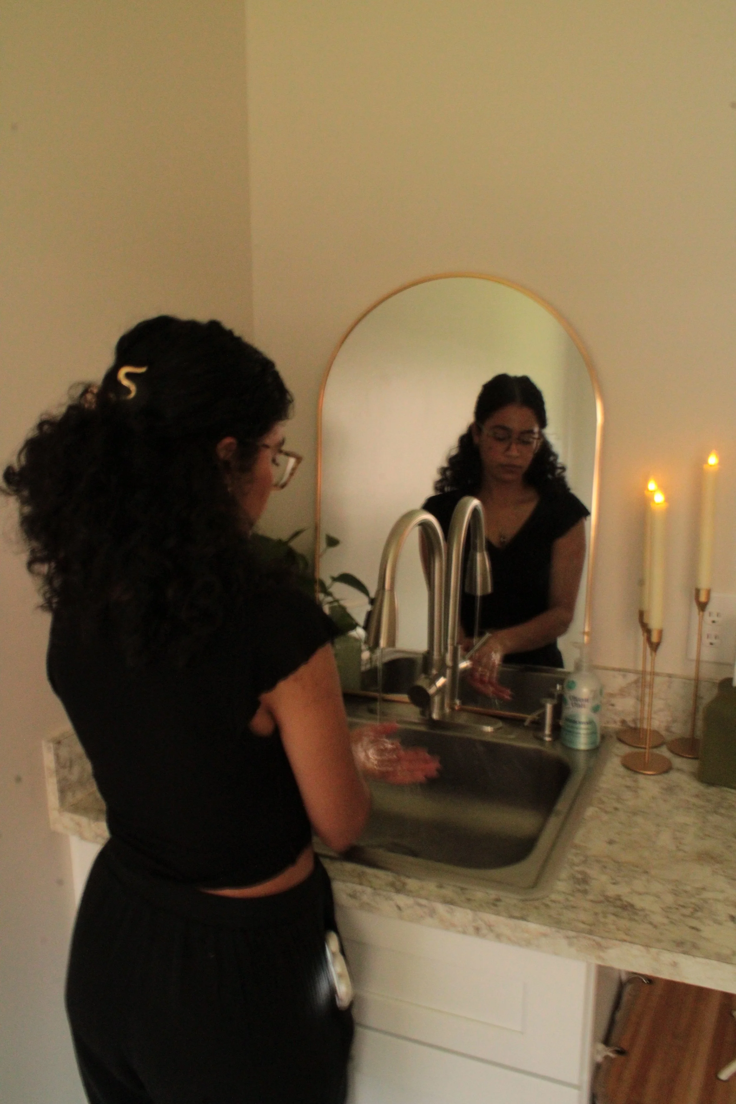 A woman with glasses and curly hair washing her hands at a kitchen sink, with a mirror reflecting her face and candles on the counter.