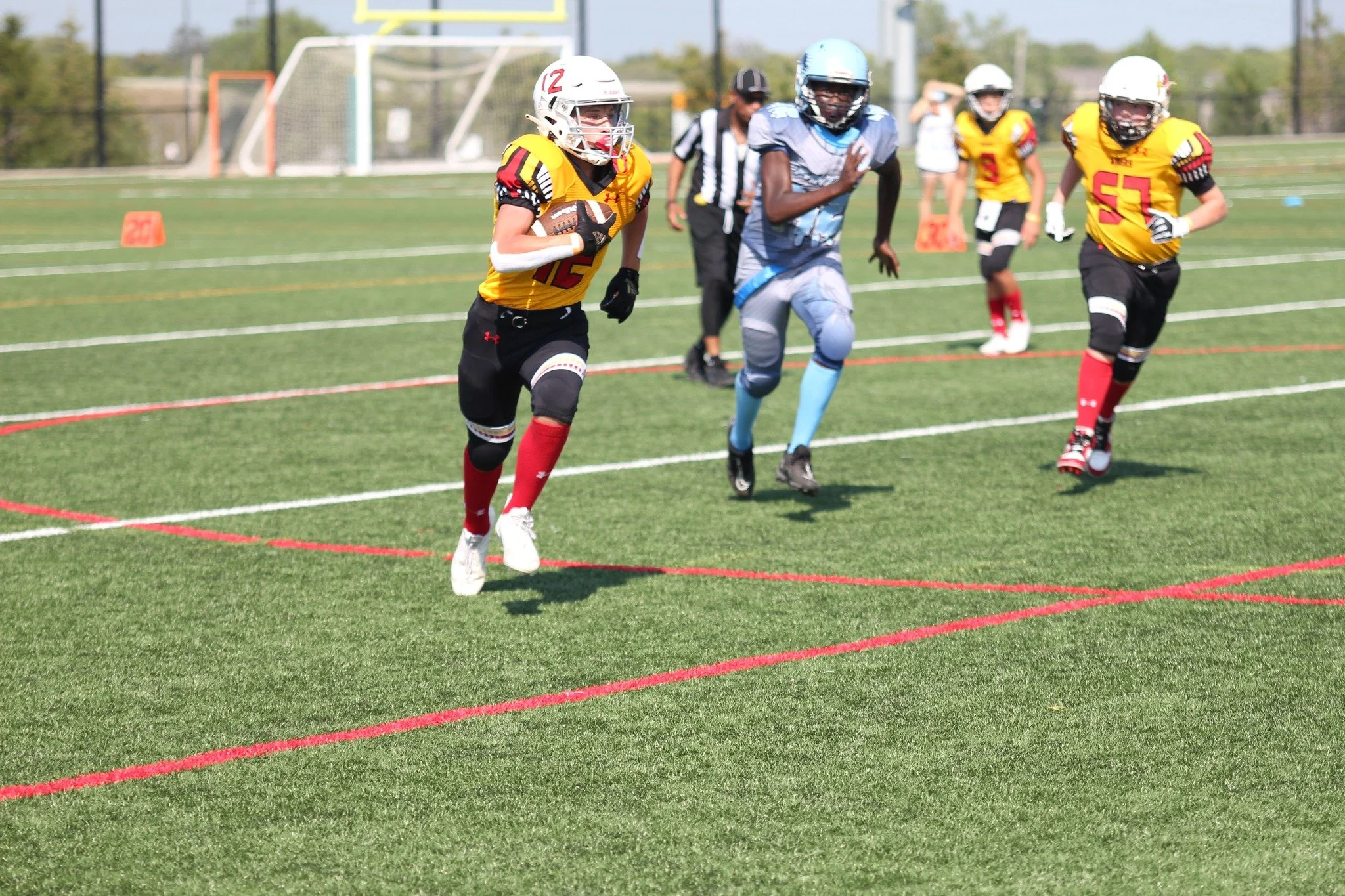 A youth football game with players running on a green field, wearing helmets and jerseys. One player in a yellow and black uniform carries the ball, while others chase behind. An official wearing a black and white striped shirt is in the background.