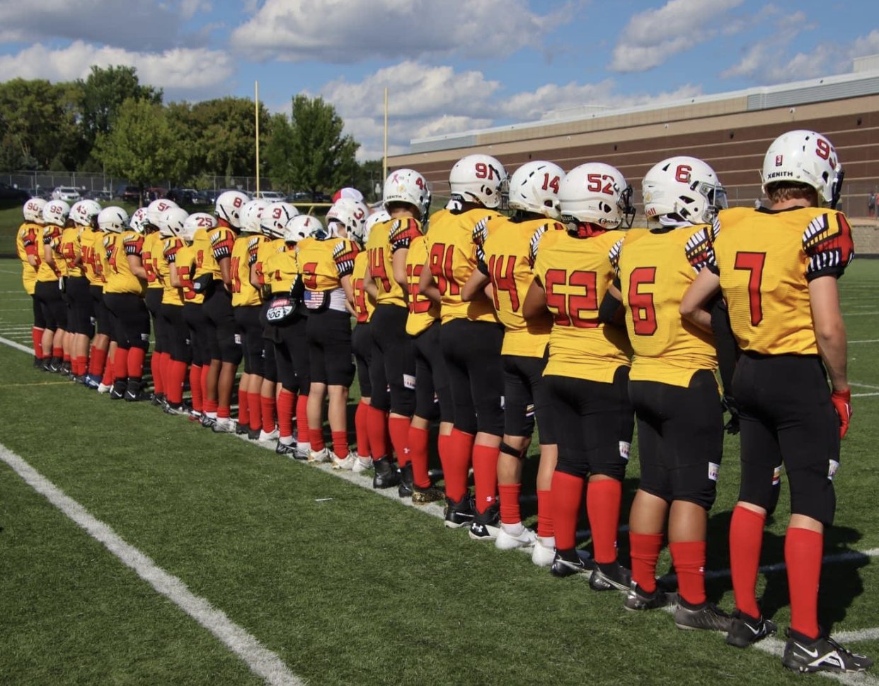 Youth football team dressed in yellow jerseys with red numbers and black pants, standing in a line on a football field.