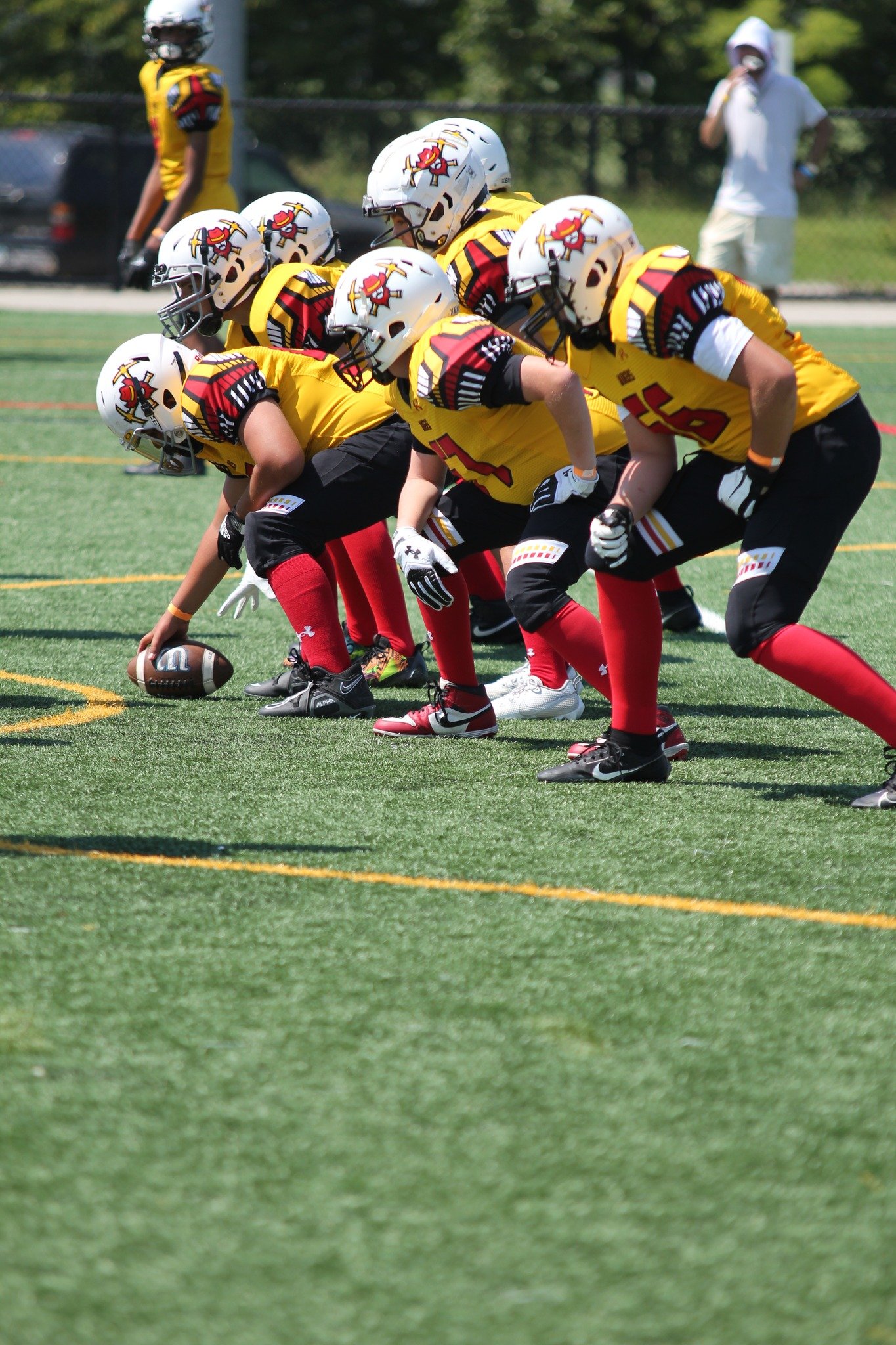 Youth football players in yellow jerseys and white helmets lined up on the field, with one player ready to snap the ball.