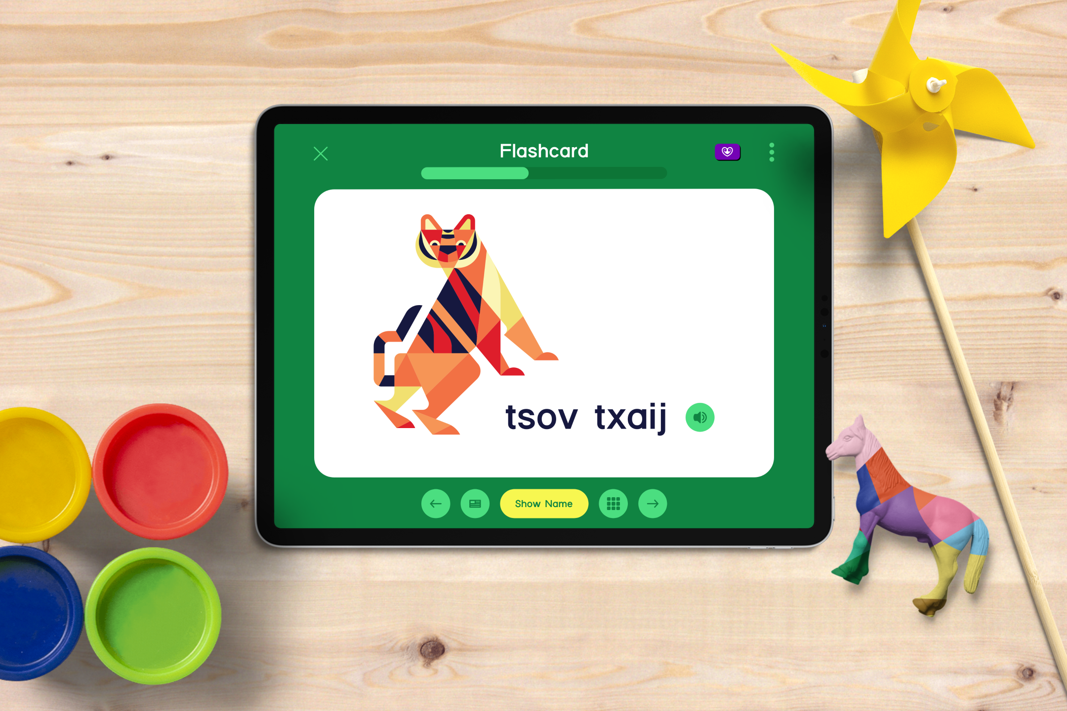 Tablet displaying an educational flashcard with a colorful geometric illustration of a tiger and the words "tsov txaj" in the center. Surrounding the tablet are small containers of paint in red, yellow, green, and blue, a yellow paper pinwheel, and a multicolored paper-mâché horse on a wooden surface.