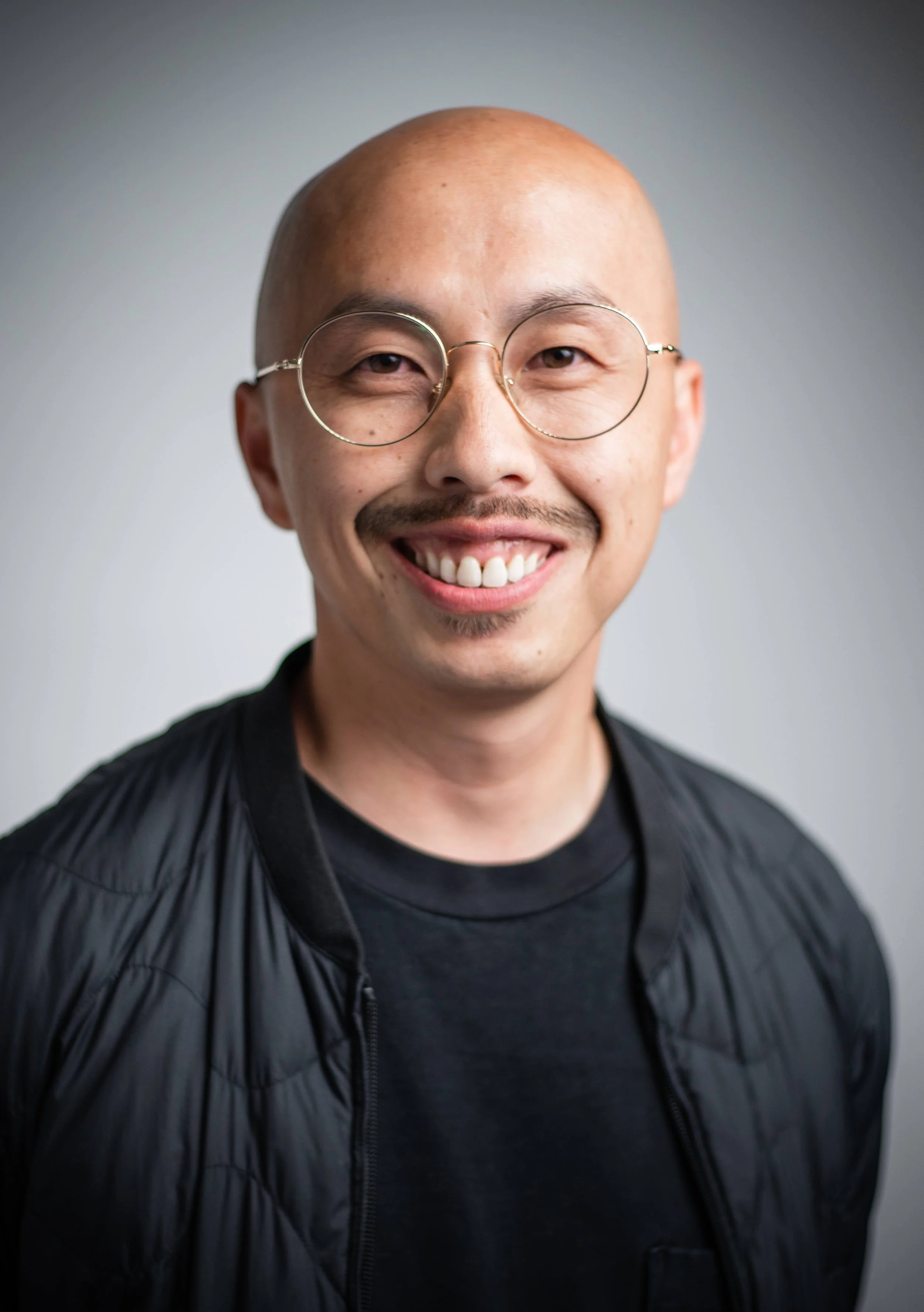 Smiling man with glasses, bald head, mustache, wearing a black jacket and t-shirt, against a plain background.