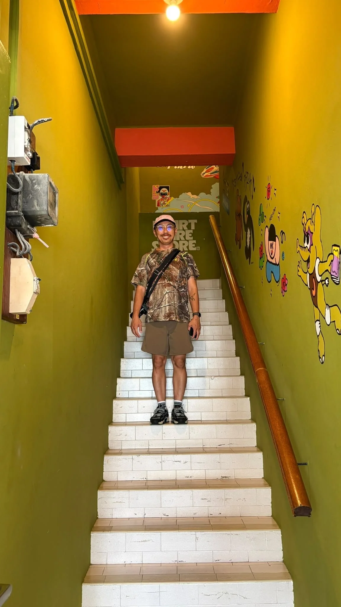 A man wearing glasses, a pink cap, camouflage shirt, beige shorts, and sneakers standing on white stairs in a colorful, cartoon-themed indoor location.