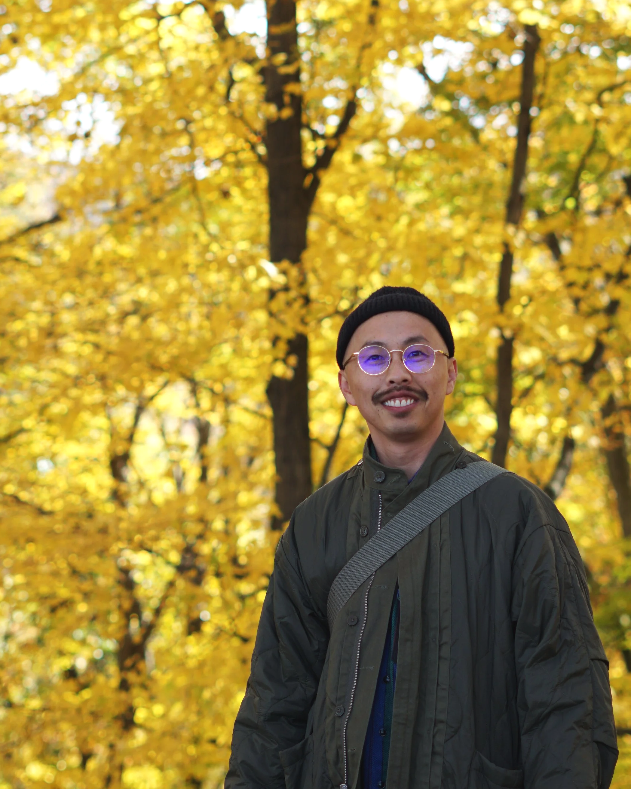 A man with a black beanie, round glasses, and a smile, standing outdoors in front of yellow autumn trees.