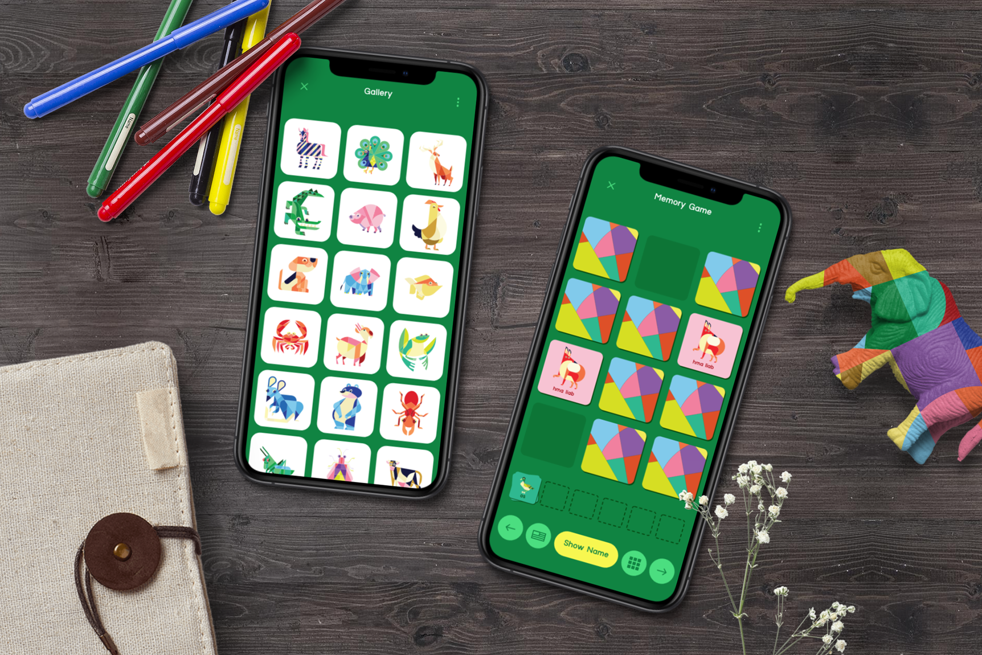 Two smartphones on a wooden table displaying colorful puzzle and matching games, surrounded by markers, a fabric notebook, a small plush dinosaur, and a sprig of white flowers.