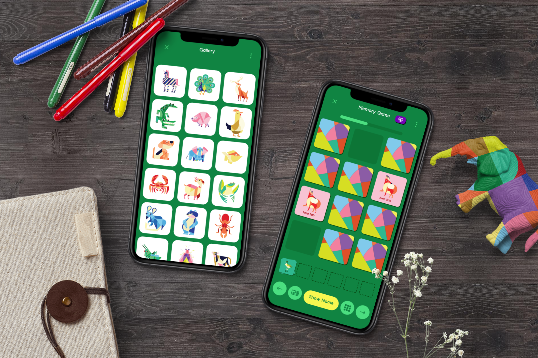 Two smartphones on a wooden table displaying digital games, with colored markers, a fabric notebook, a colorful fabric elephant, and small white flowers nearby.