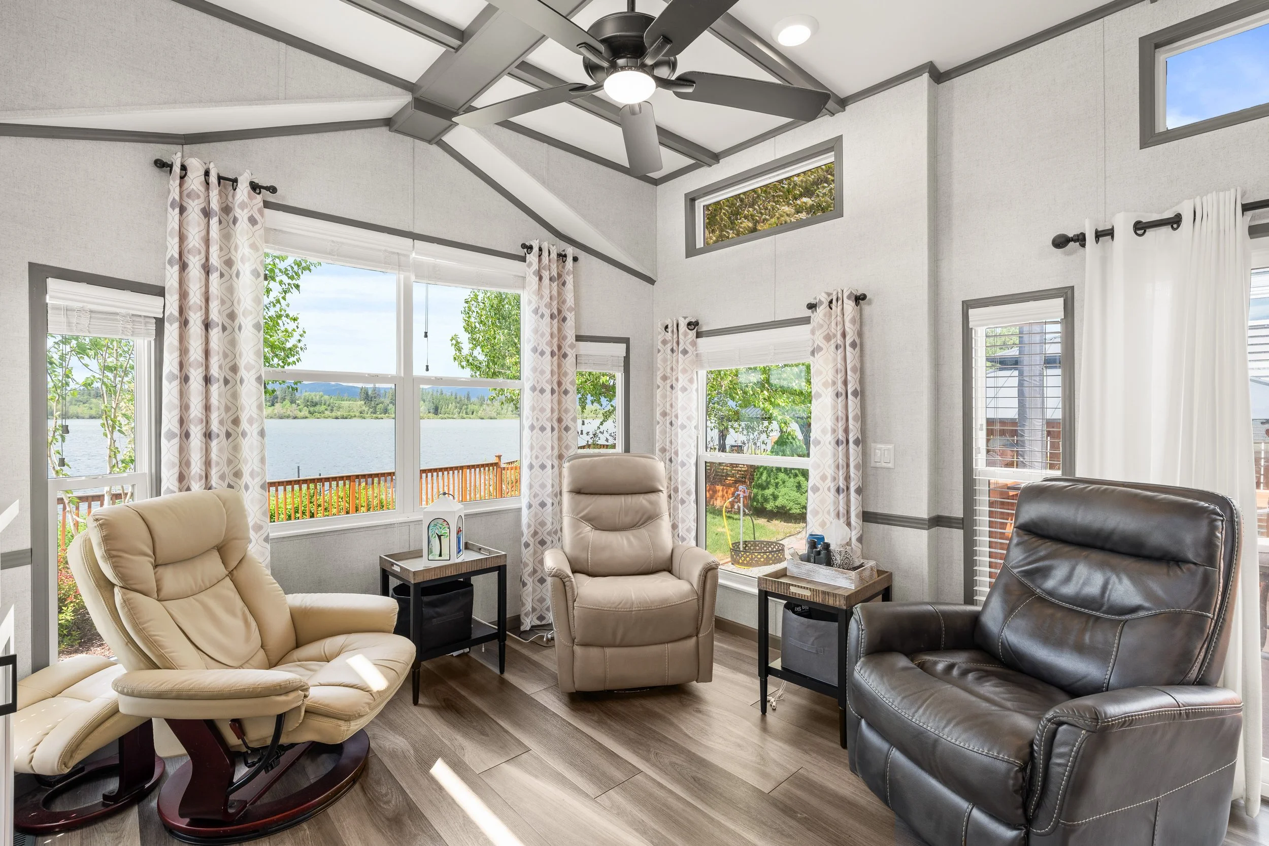 Pend Oreille County Real Estate Photography - Living room with large windows overlooking water, featuring beige and dark leather armchairs, curtains, small side tables, and a ceiling fan.