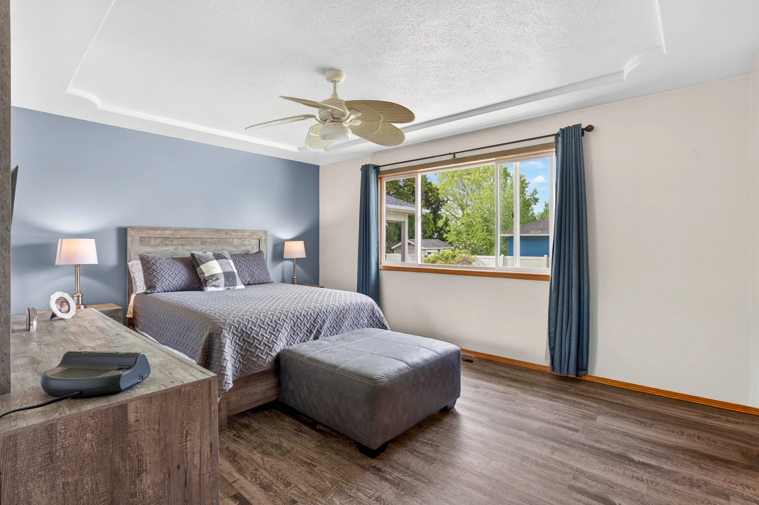 Spokane Real Estate Photography - A bedroom with a large window with blue curtains, a bed with a patterned gray bedspread and pillows, two nightstands with lamps, a ceiling fan, and a wooden table with some items on it, including a black speaker.