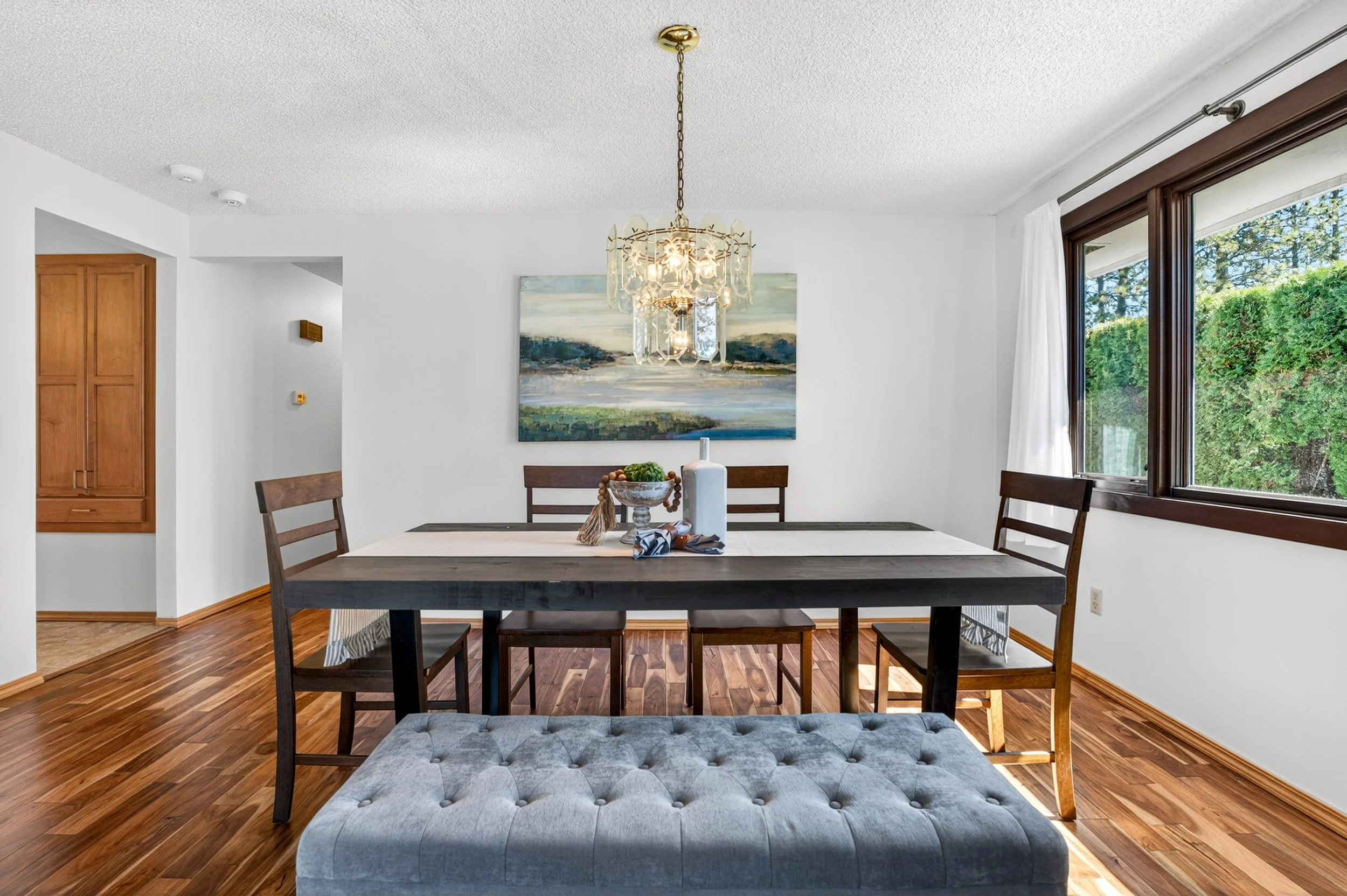 Spokane Real Estate Photography - A dining room with a rectangular table, six chairs, a hanging chandelier, a large window with white curtains, and a landscape painting on the wall.