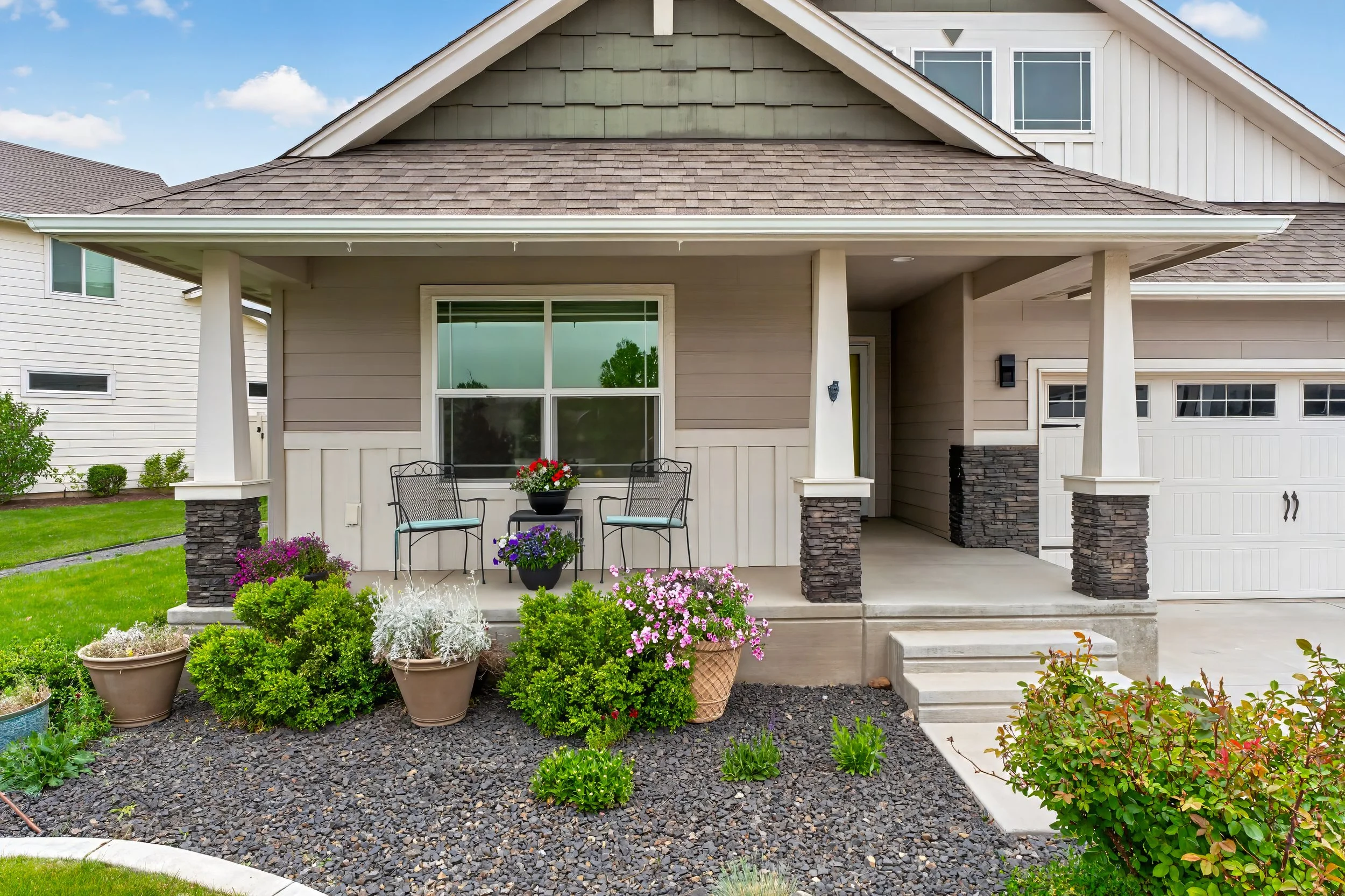 Spokane Real Estate Photography - A suburban house with a porch and garden. The porch has two chairs and a small table with a flowerpot. The garden features various plants and flowers, with a rock mulch ground cover.