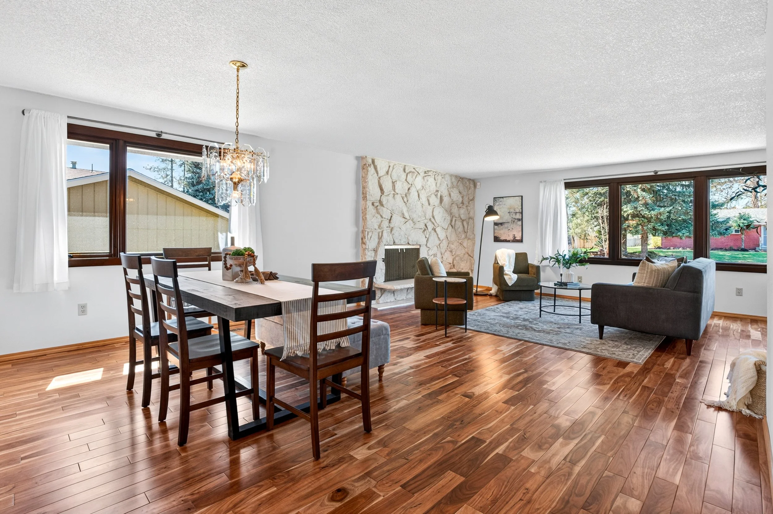 Spokane Real Estate Photography - Living room with large windows, hardwood floors, a dining table with chairs, a stone fireplace, and seating area with chairs and a rug.