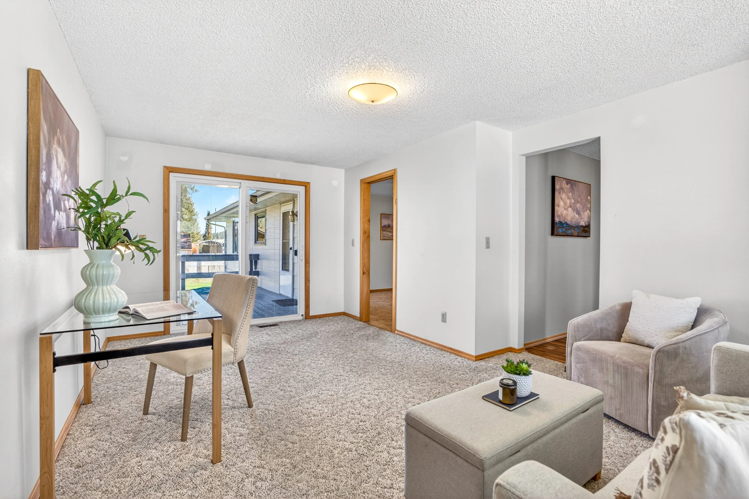 Spokane Real Estate Photography - Living room with beige carpet, white walls, a small glass desk with a beige chair, a potted plant, art on the walls, a ceiling light, and a sliding glass door leading outside.
