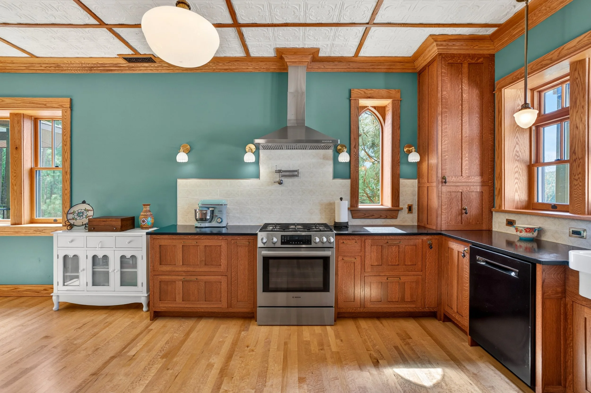 Chewelah Real Estate Photography - A kitchen with green walls, wooden cabinets, and a stainless steel oven. There's a white sideboard with glass-paneled doors, decorative items, and a window showing trees outside.