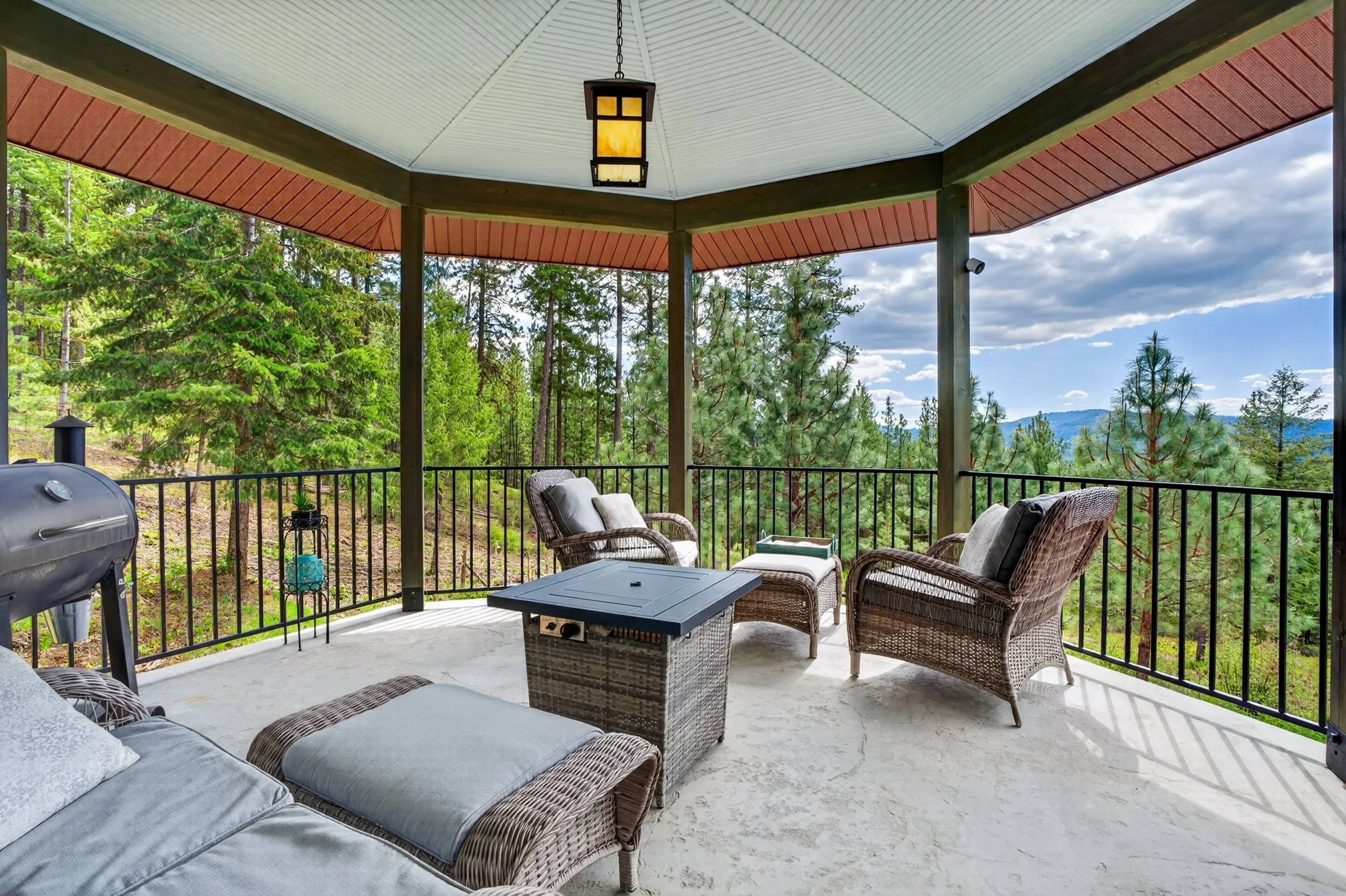 Chewelah Real Estate Photography - Covered porch overlooking lush green forest and distant mountains, furnished with wicker chairs, a gray couch, and a small black table, with a ceiling lantern and a grill on the side.