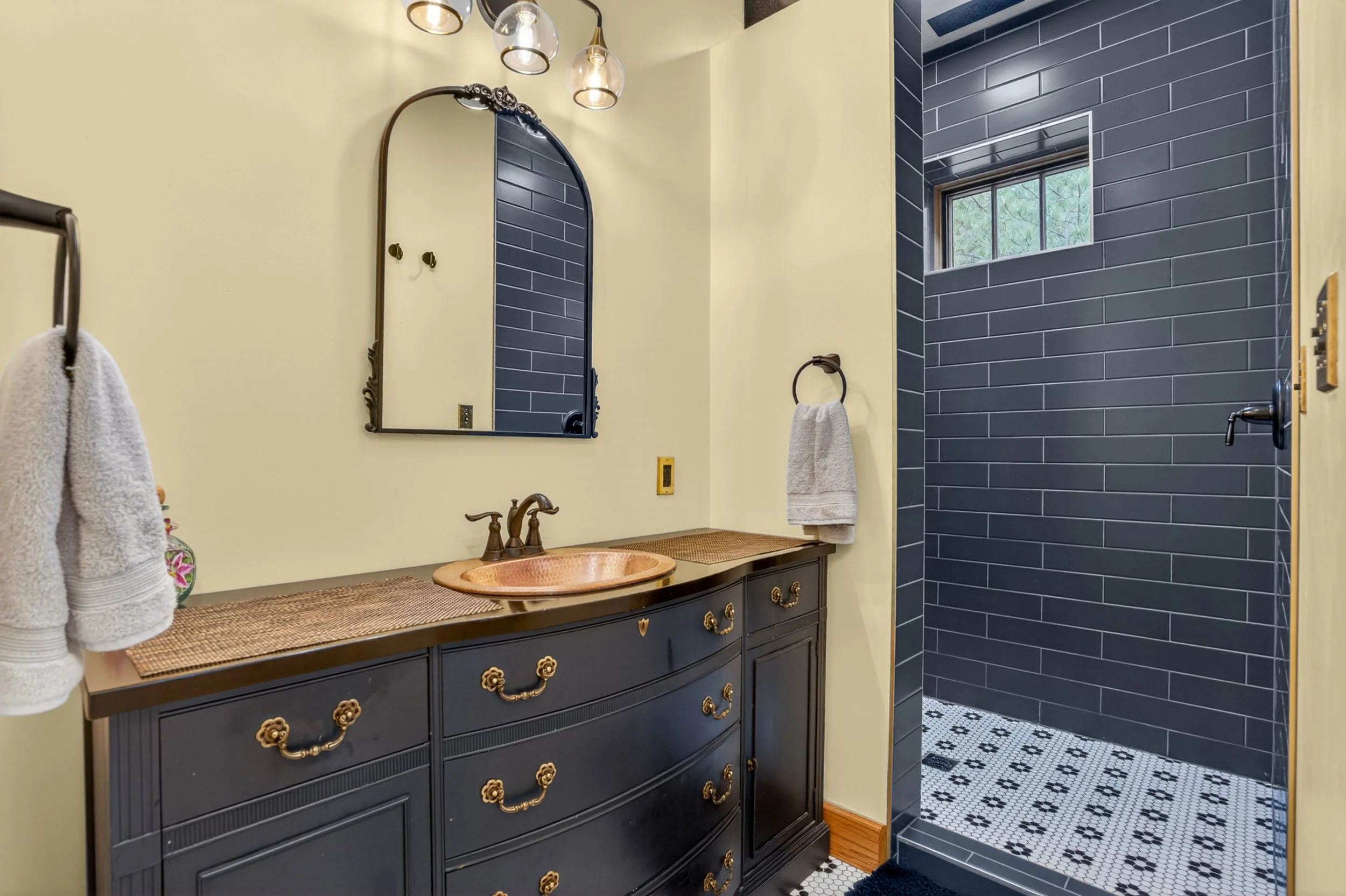 Chewelah Real Estate Photography - Bathroom with a black vanity featuring ornate gold hardware, a copper sink, and a large mirror. The wall is painted yellow, and a black towel ring holds a white towel. 