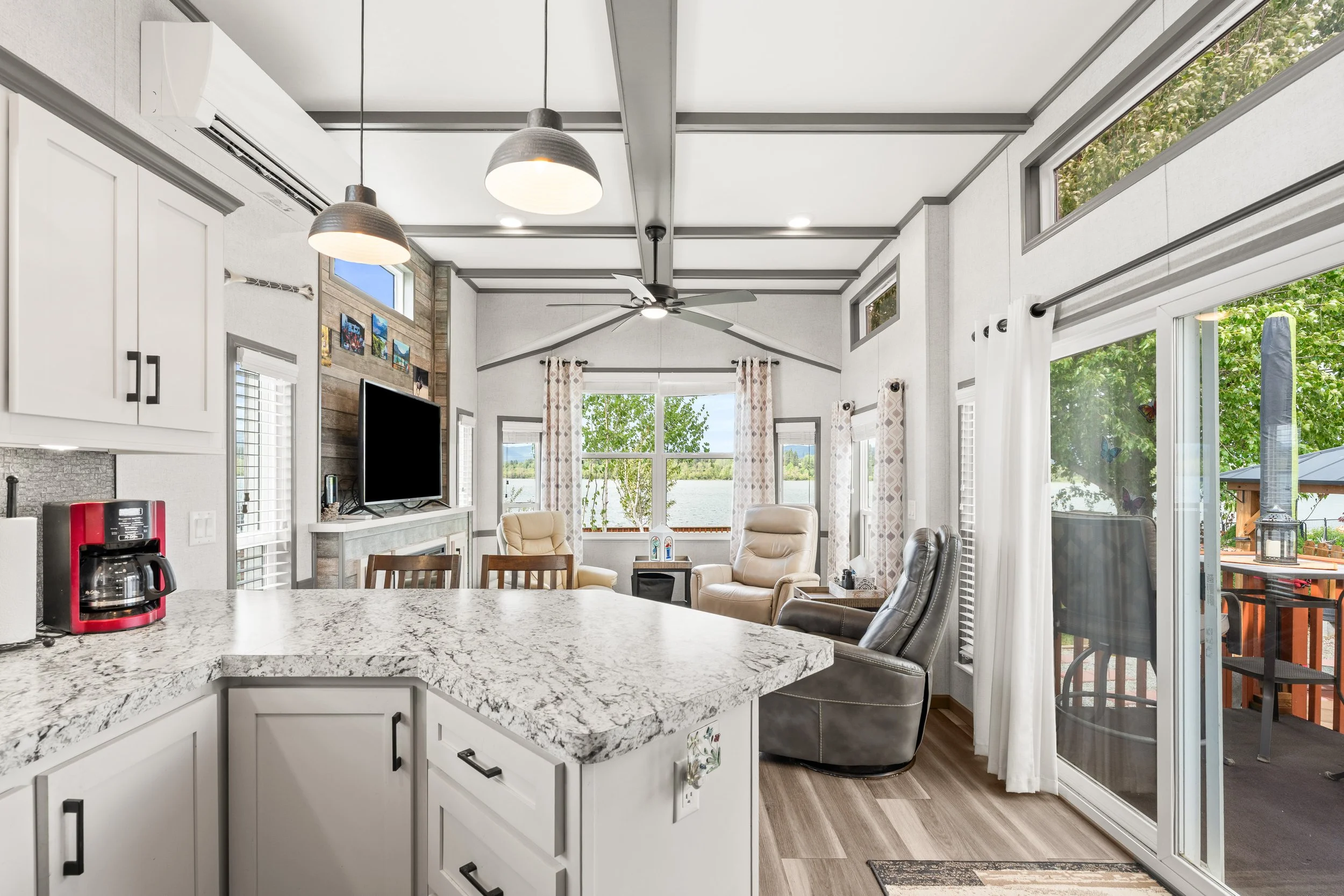 Pend Oreille County Real Estate Photography - Open-concept living room seen from kitchen with granite countertop in the foreground, white cabinets, ceiling fans, multiple windows with curtains, and a sliding glass door.