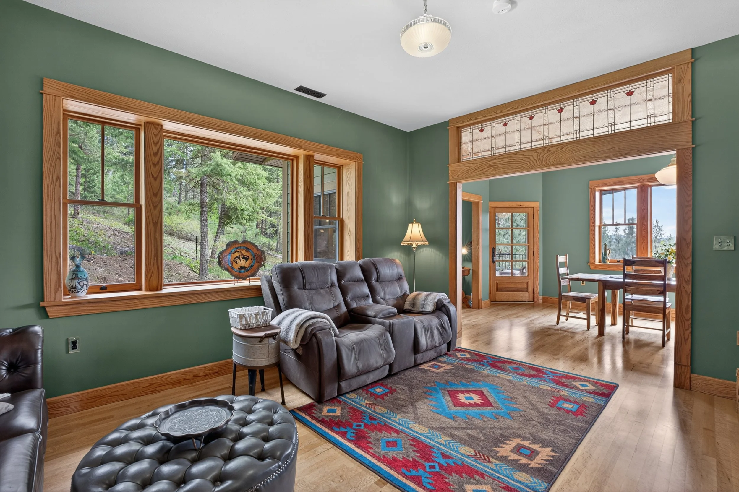 Chewelah Real Estate Photography - Living room with green walls, large windows overlooking a wooded area, and wooden trim. 