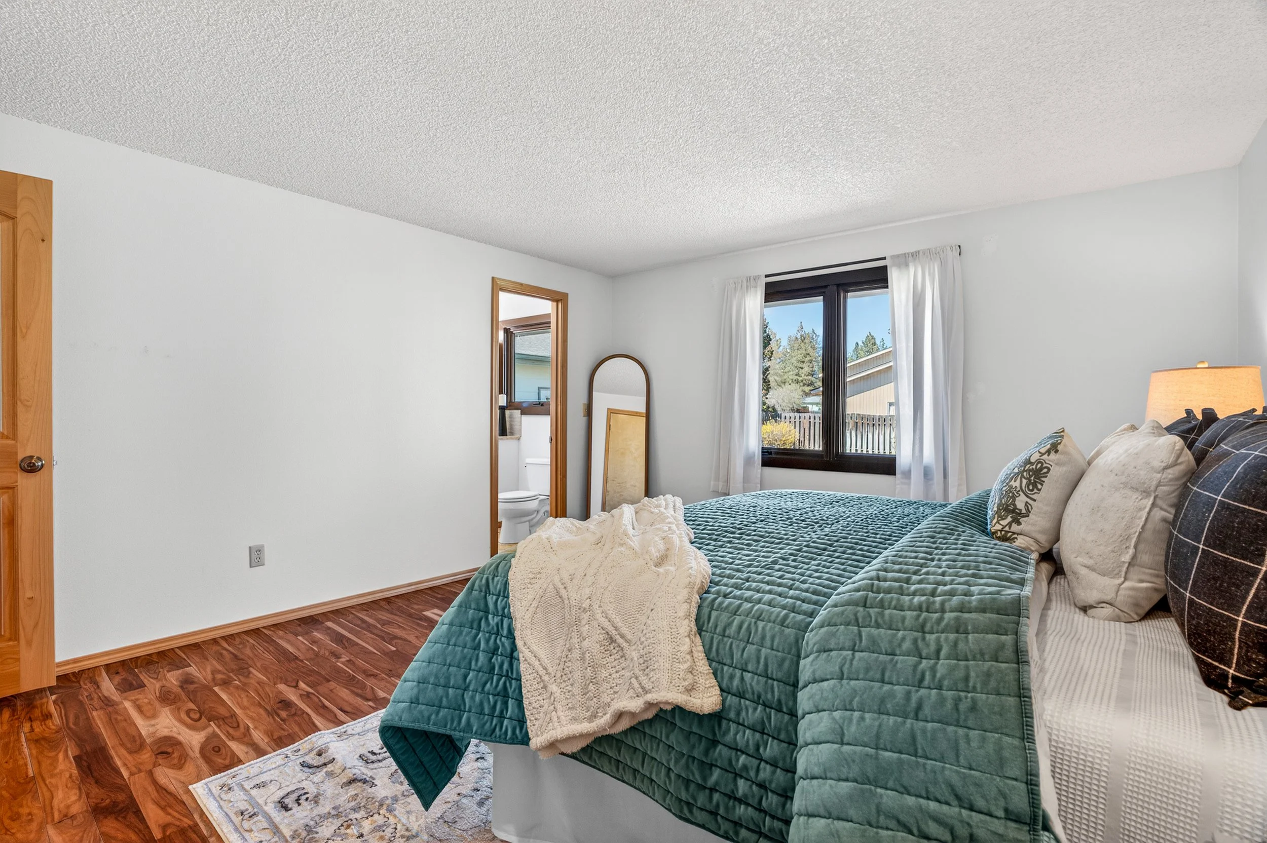 Spokane Real Estate Photography - Bedroom with white walls, wooden flooring, bed with green quilt and multiple pillows, window with white curtains, mirror leaning against wall, and a glimpse of bathroom visible through door.