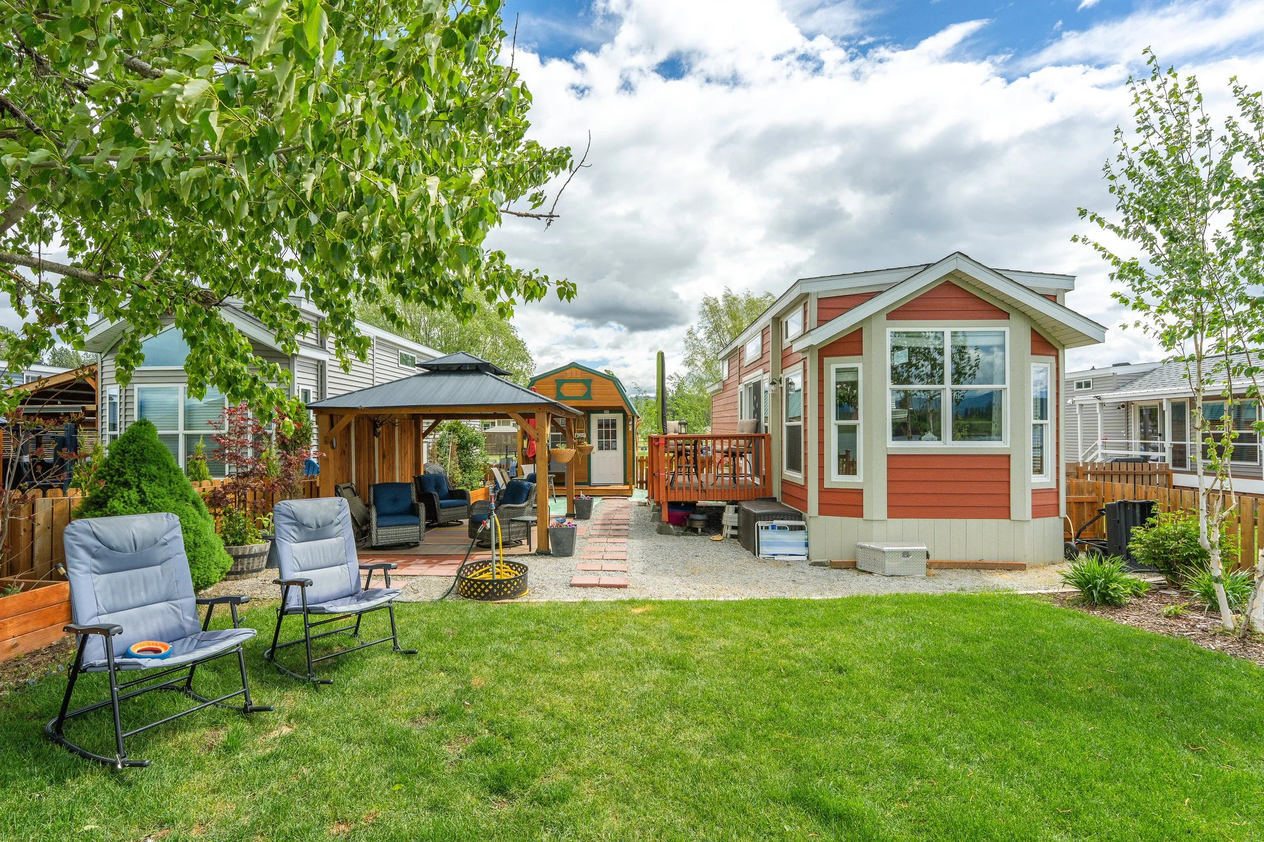 Pend Oreille County Real Estate Photography - Backyard patio with chairs, a small table, a wooden gazebo, a red house with white trim, various potted plants, and neighboring houses under a partly cloudy sky.