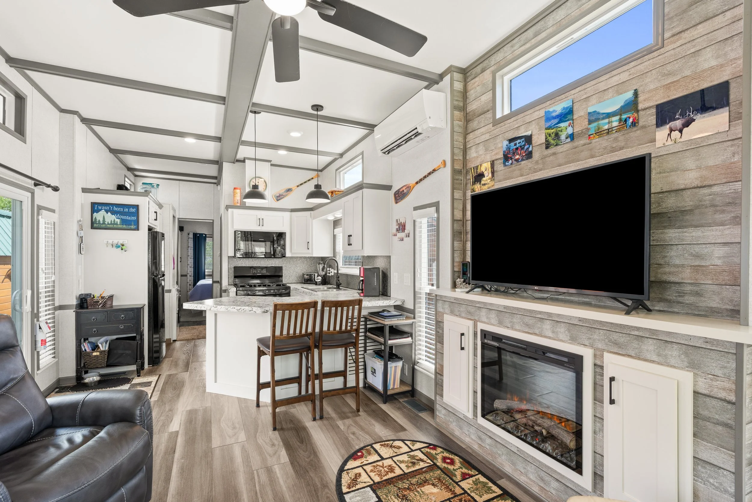 Pend Oreille County Real Estate Photography - Interior view of a living space with a partial view of a kitchen. The room features high ceilings with exposed beams, a stone accent wall with a mounted flat-screen TV, and a fireplace below it. 