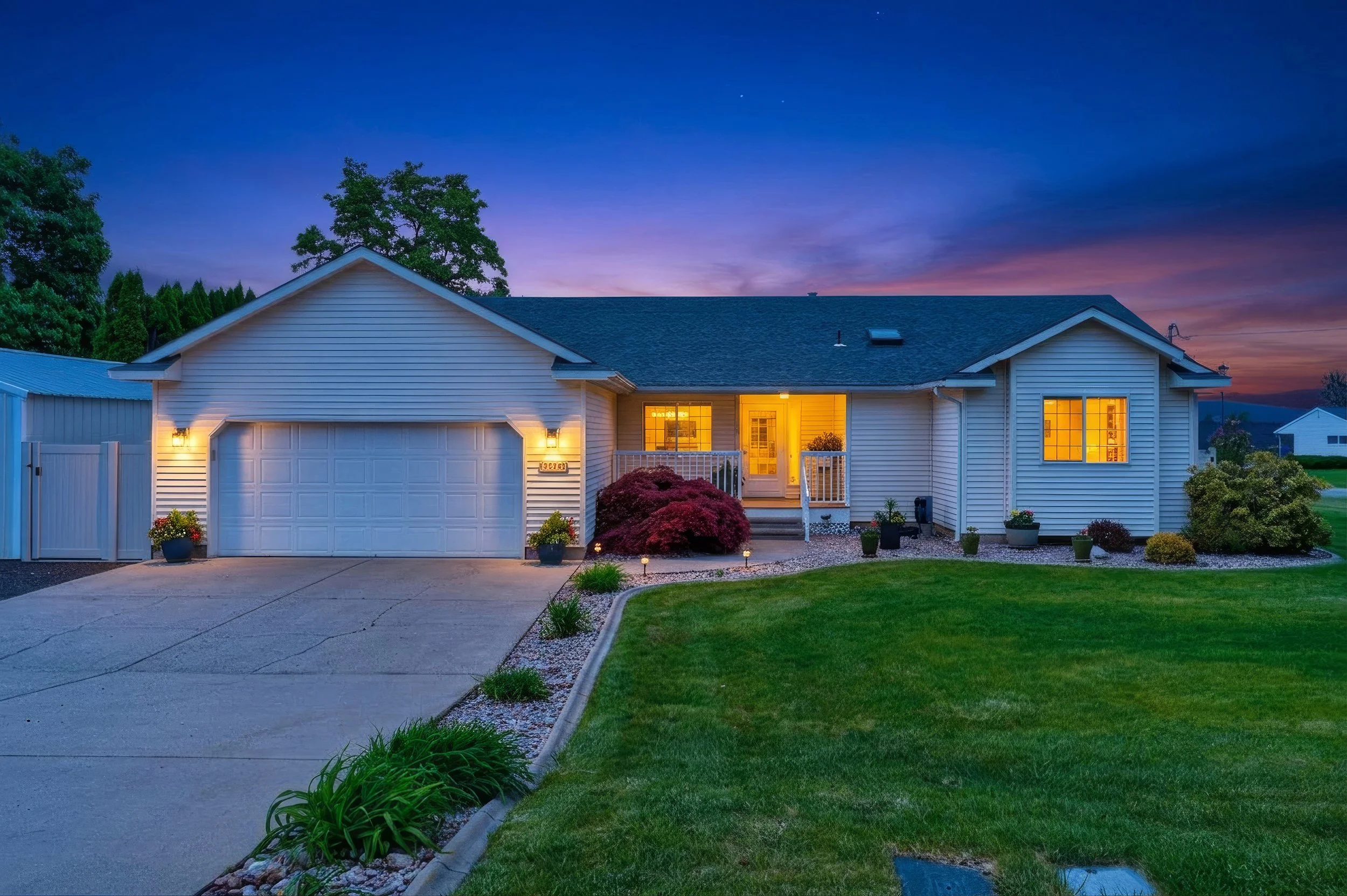 Spokane Real Estate Photography - Virtual twilight image of a house with lit windows at dusk, with a well-maintained front yard, a driveway, and trees in the background.