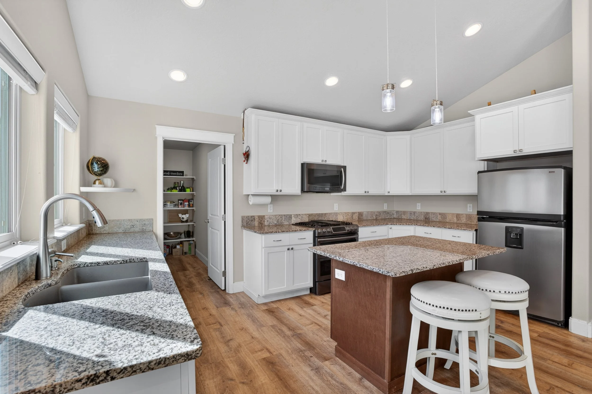 Spokane Real Estate Photography - View of a modern kitchen with white cabinets, granite countertops, hardwood floors, stainless steel appliances, and a kitchen island with two white stools.