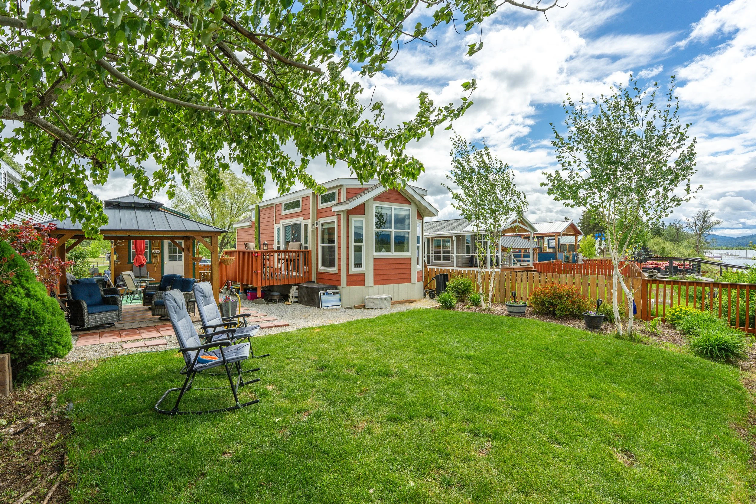 Pend Oreille County Real Estate Photography - A backyard with green grass, patio furniture, a wooden deck, and a small house with large windows, surrounded by trees and a fenced yard under a partly cloudy sky.