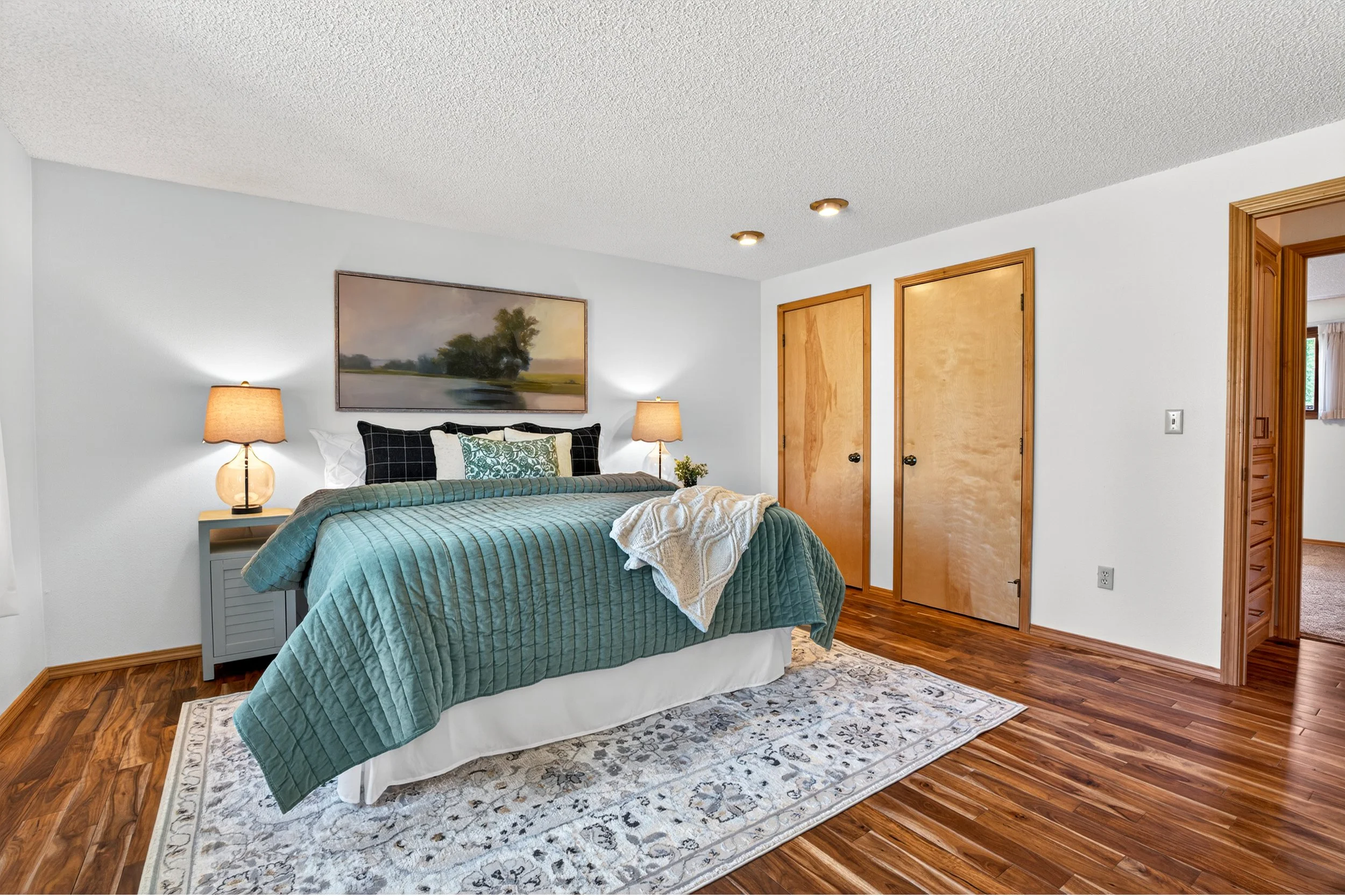 Spokane Real Estate Photography - A bedroom with a bed covered in a teal quilt, two bedside lamps, a framed landscape painting above the bed, and wooden closet doors, with a patterned rug on hardwood flooring.